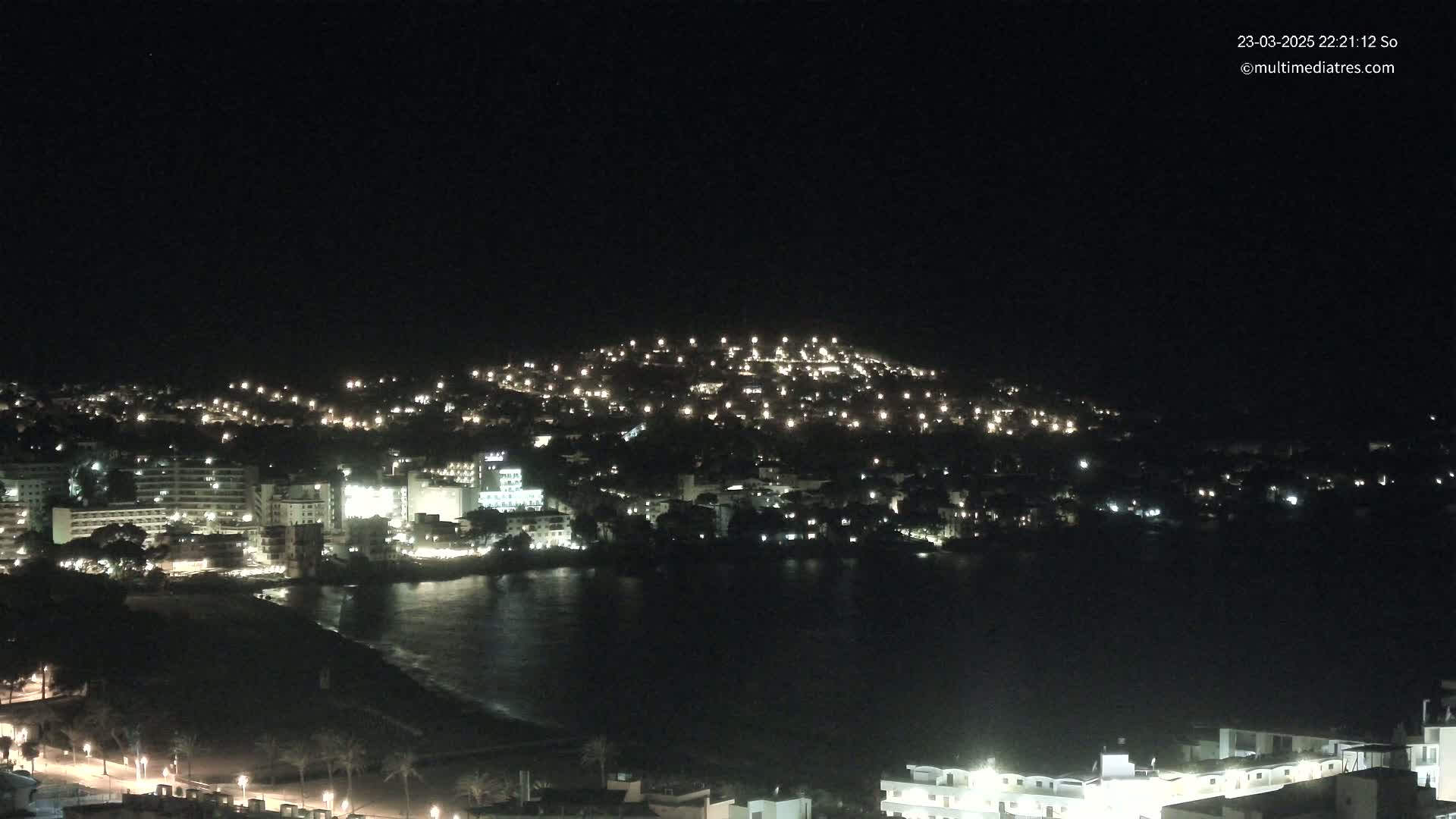 A nighttime view of a coastal town illuminated by many lights, with a dark body of water in front and a hill rising behind it.