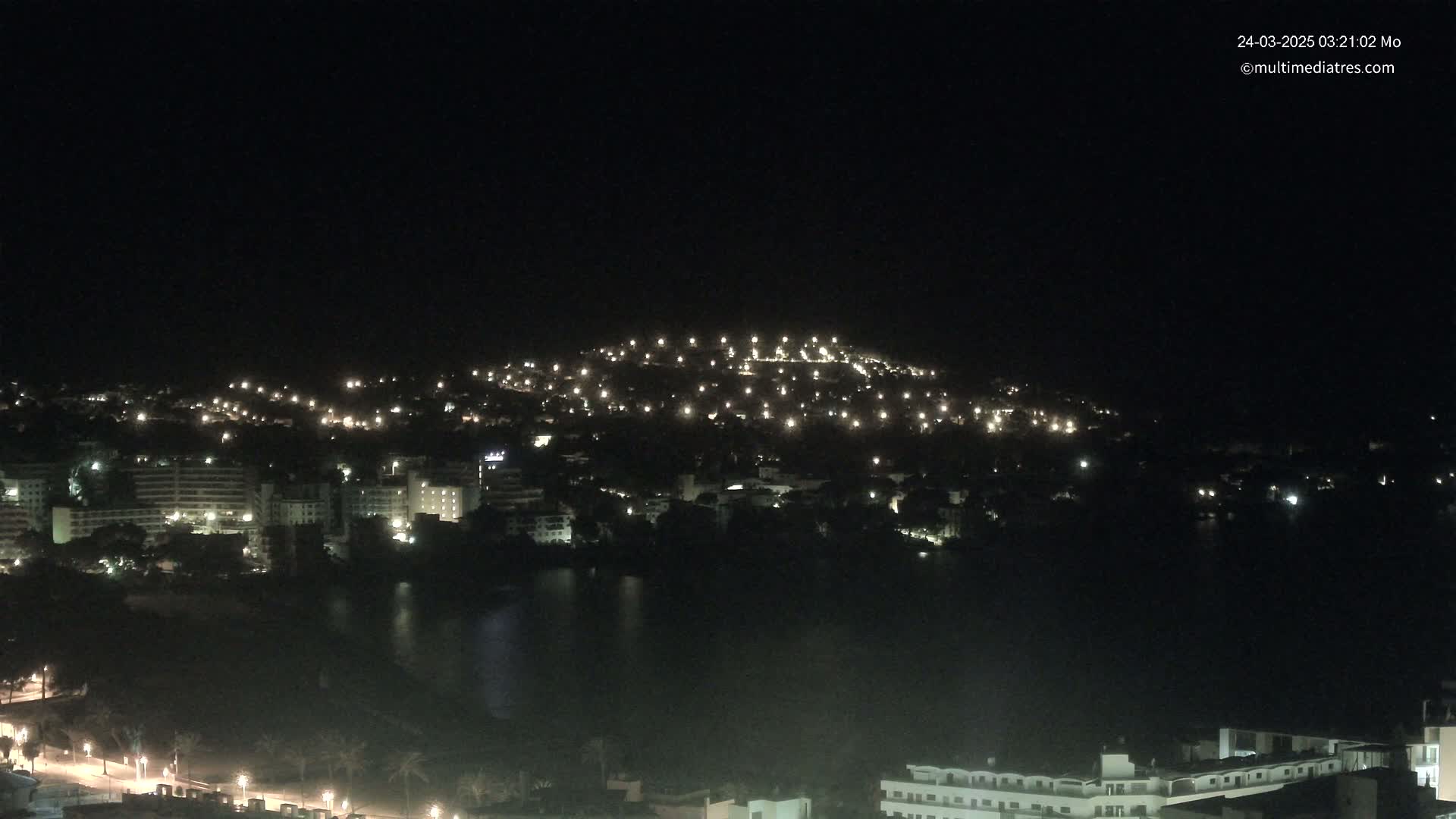 A nighttime view of a coastal town with numerous lights illuminating buildings and streets, situated around a dark body of water under a clear, dark sky.