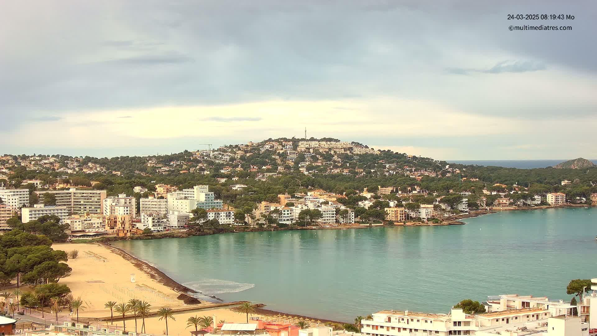 A coastal town with numerous buildings and a sandy beach is situated on a bay under a mostly cloudy sky.