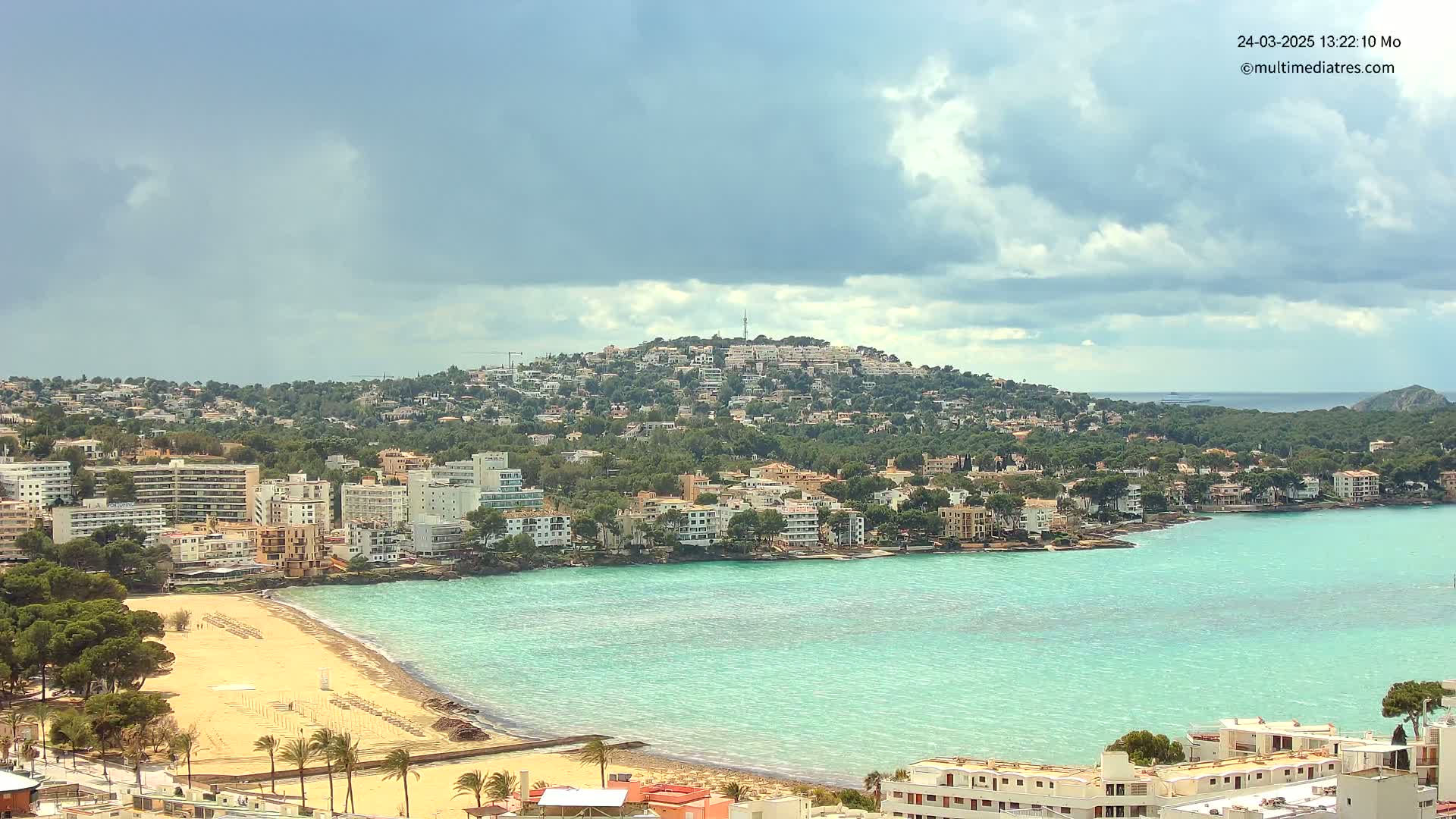 A coastal town with a sandy beach and turquoise water is nestled against a hill covered in buildings and trees under a partly cloudy sky.