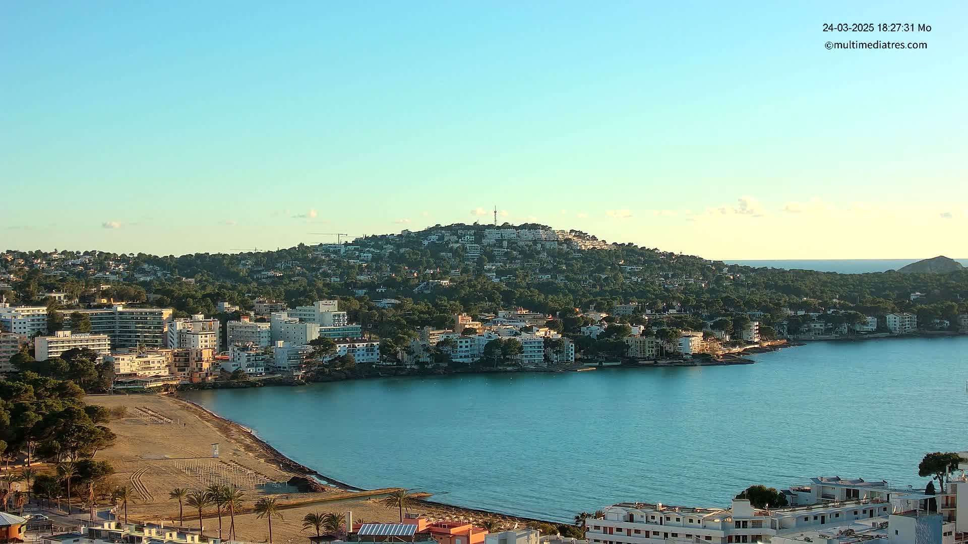 A coastal town nestled against a hill, with a sandy beach and calm, blue water under a clear sky.