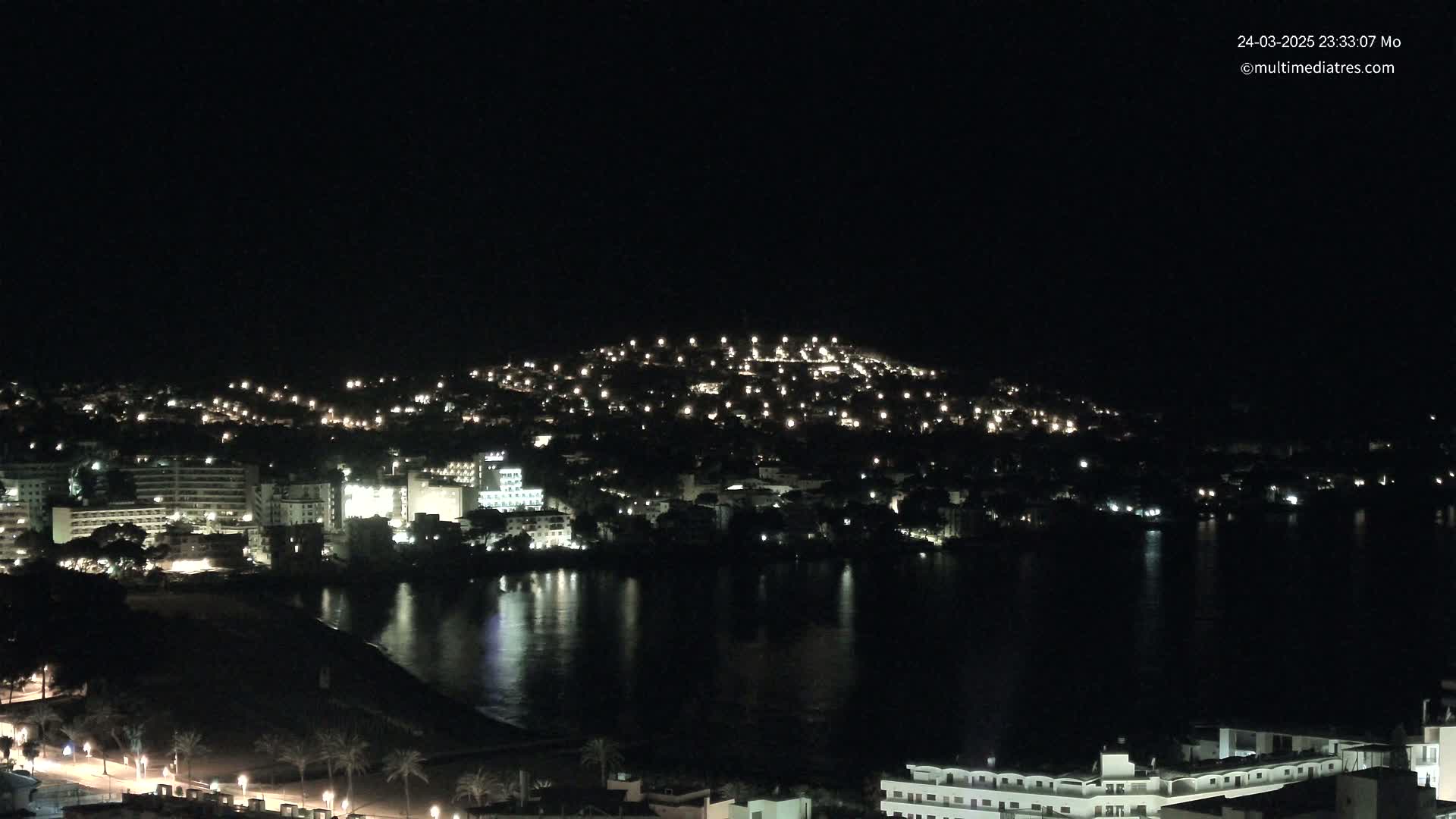 A nighttime view of a coastal town illuminated by many lights, bordered by dark water reflecting the lights.