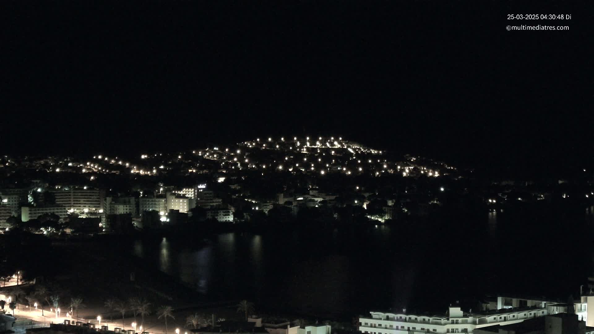 A nighttime view of a city with numerous lights illuminating buildings and streets, situated beside a dark body of water under a clear, dark sky.