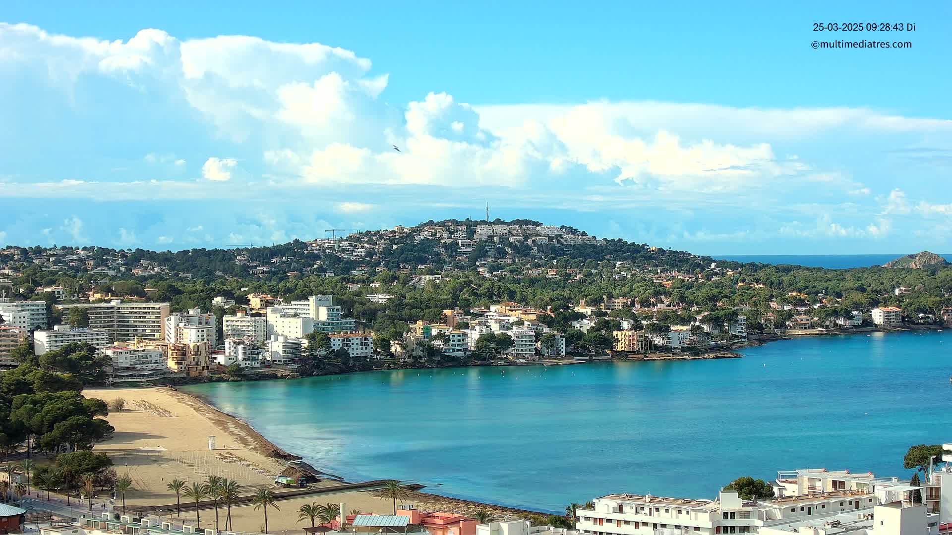 A coastal town nestled beside a calm, turquoise bay under a partly cloudy blue sky.