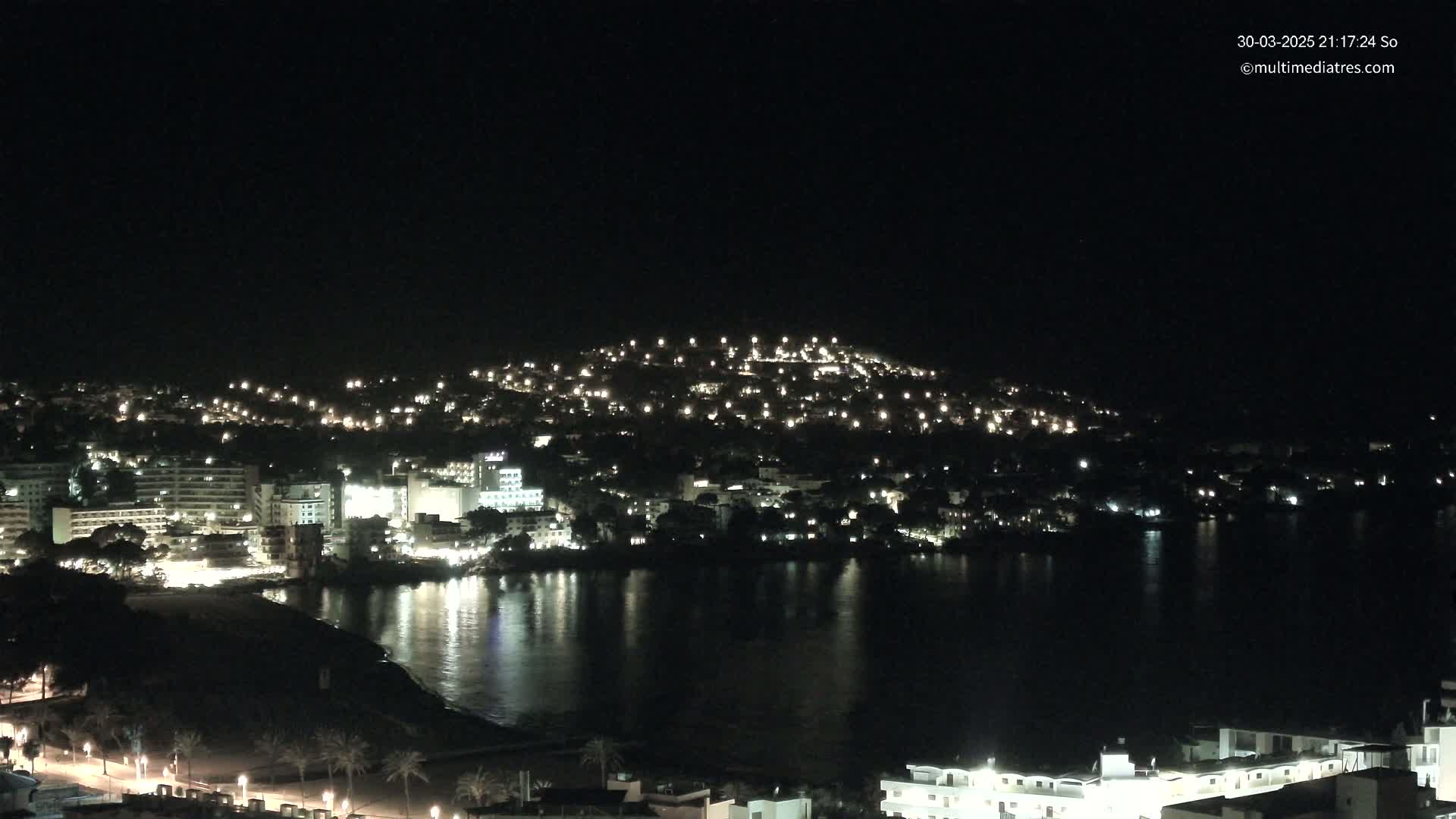 A nighttime view of a coastal town illuminated by many lights, with a calm body of water reflecting the lights in the foreground.