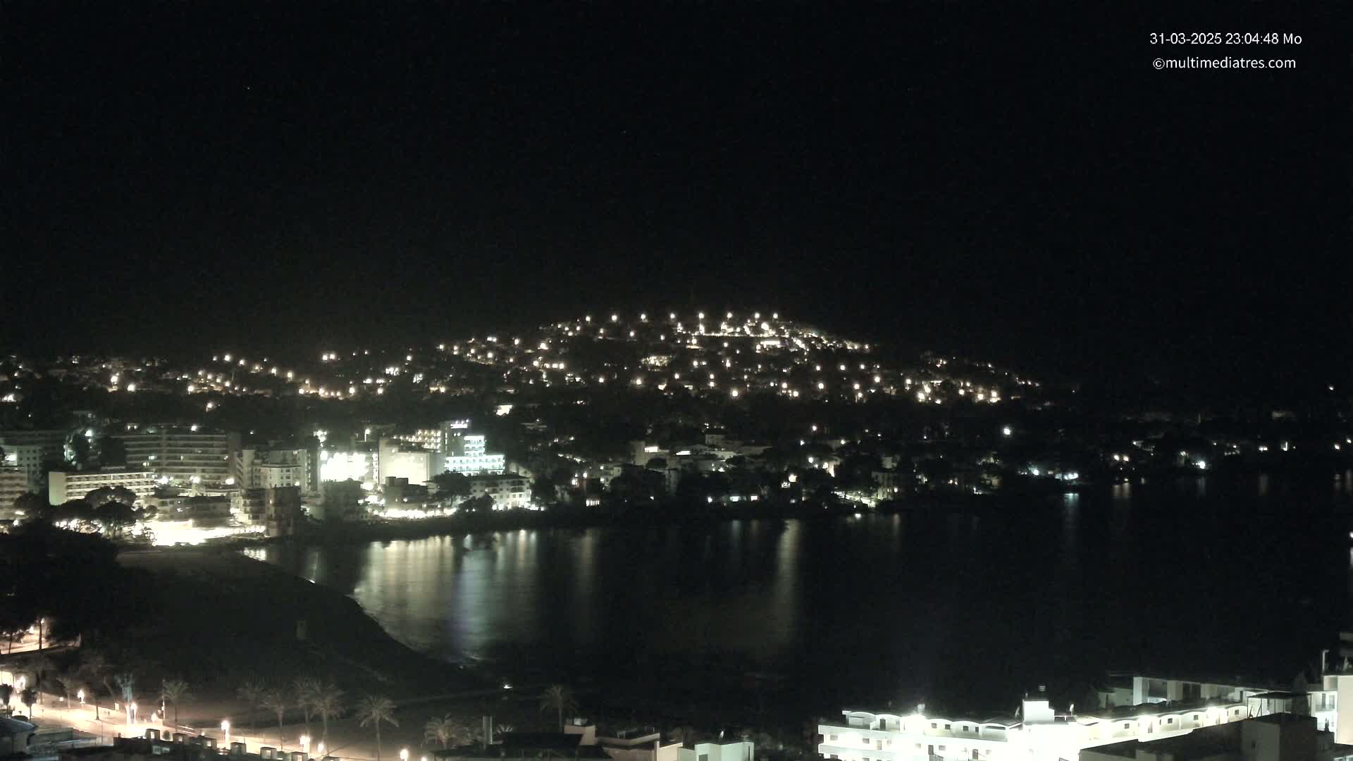 A nighttime view of a coastal town with numerous lights illuminating buildings and a calm body of water.