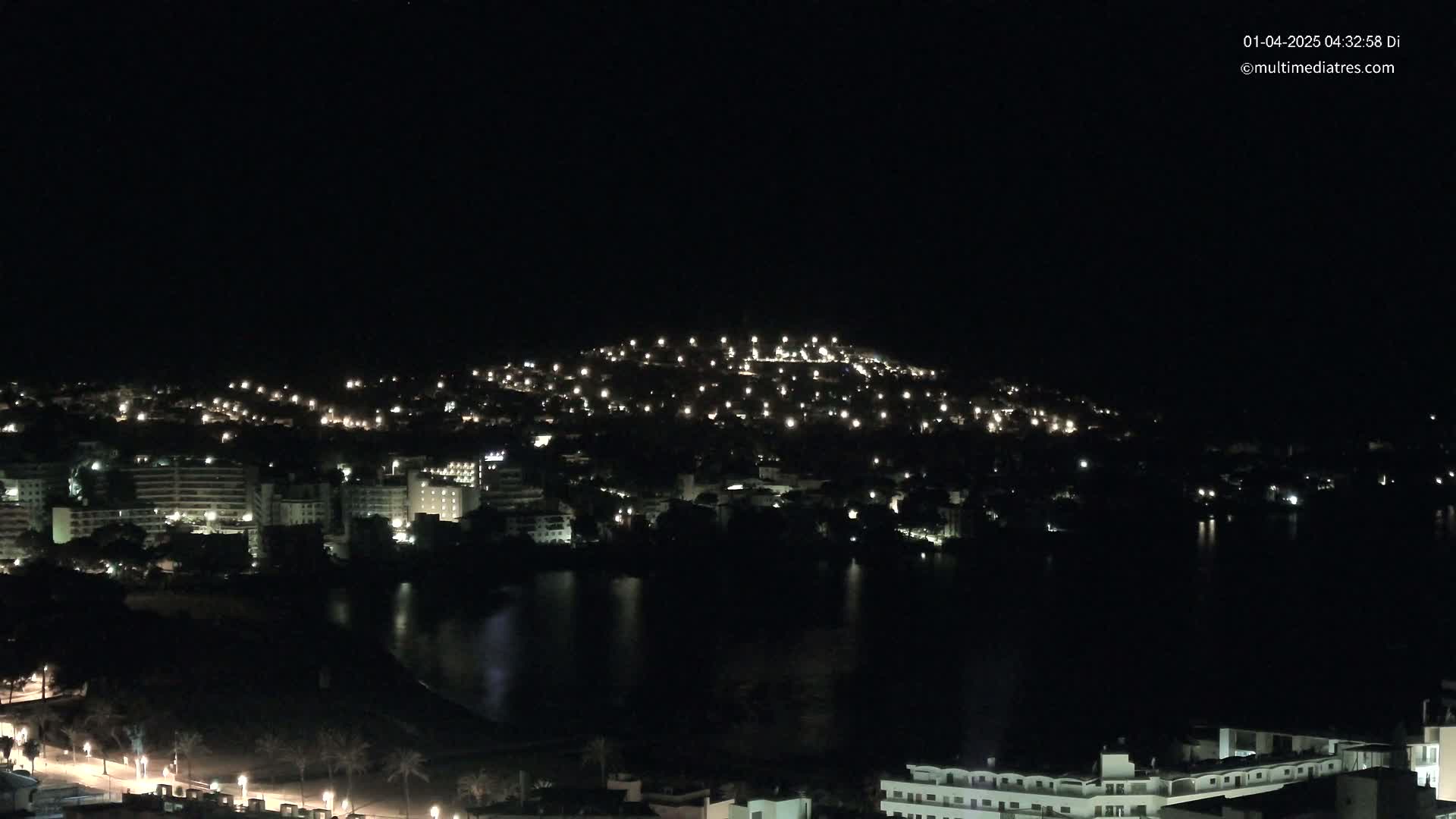 A nighttime view of a coastal town, illuminated by scattered lights, with a dark body of water in the foreground.
