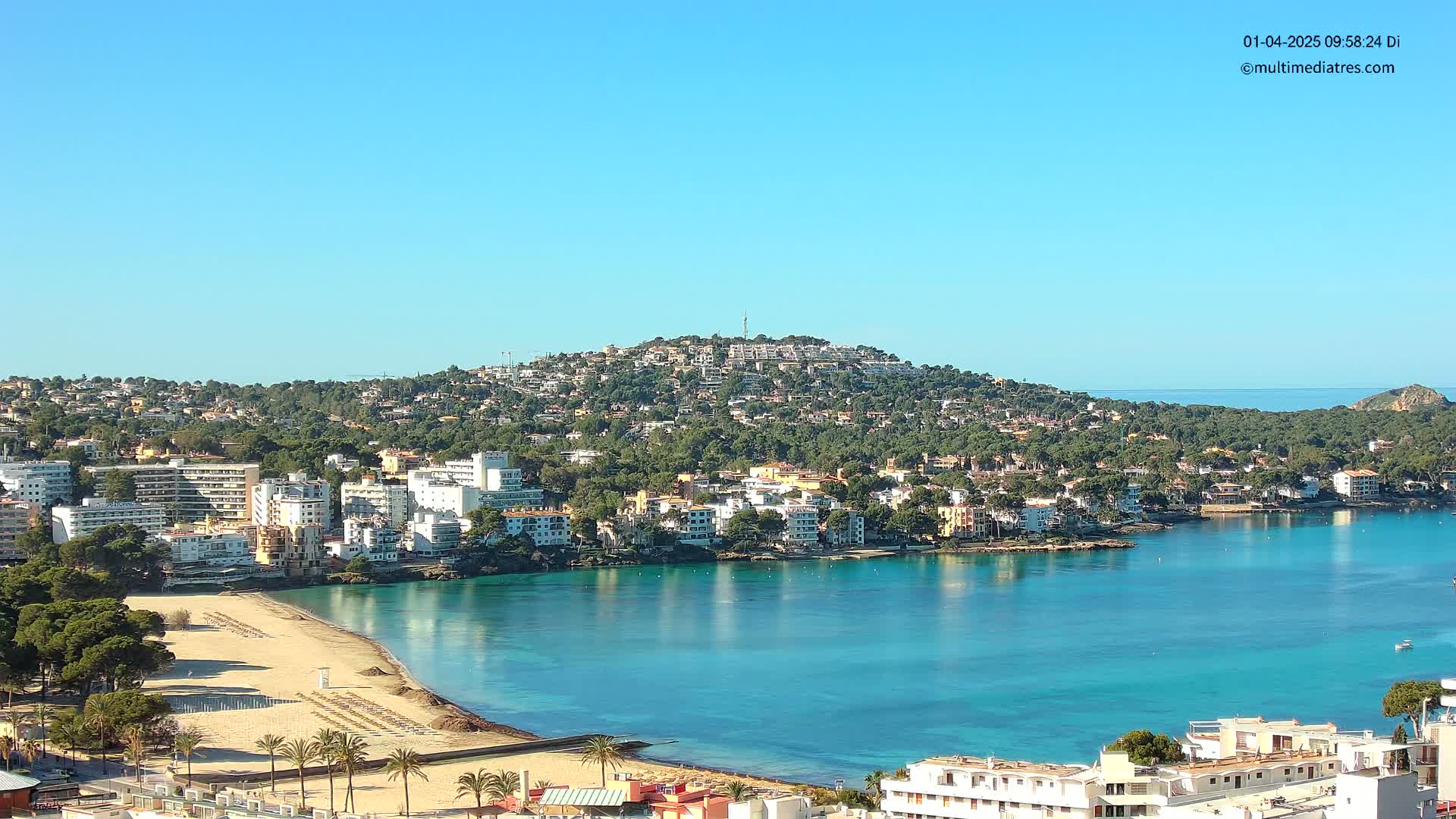 A coastal town nestled against a verdant hill, with a calm turquoise bay and sandy beach, under a clear blue sky.