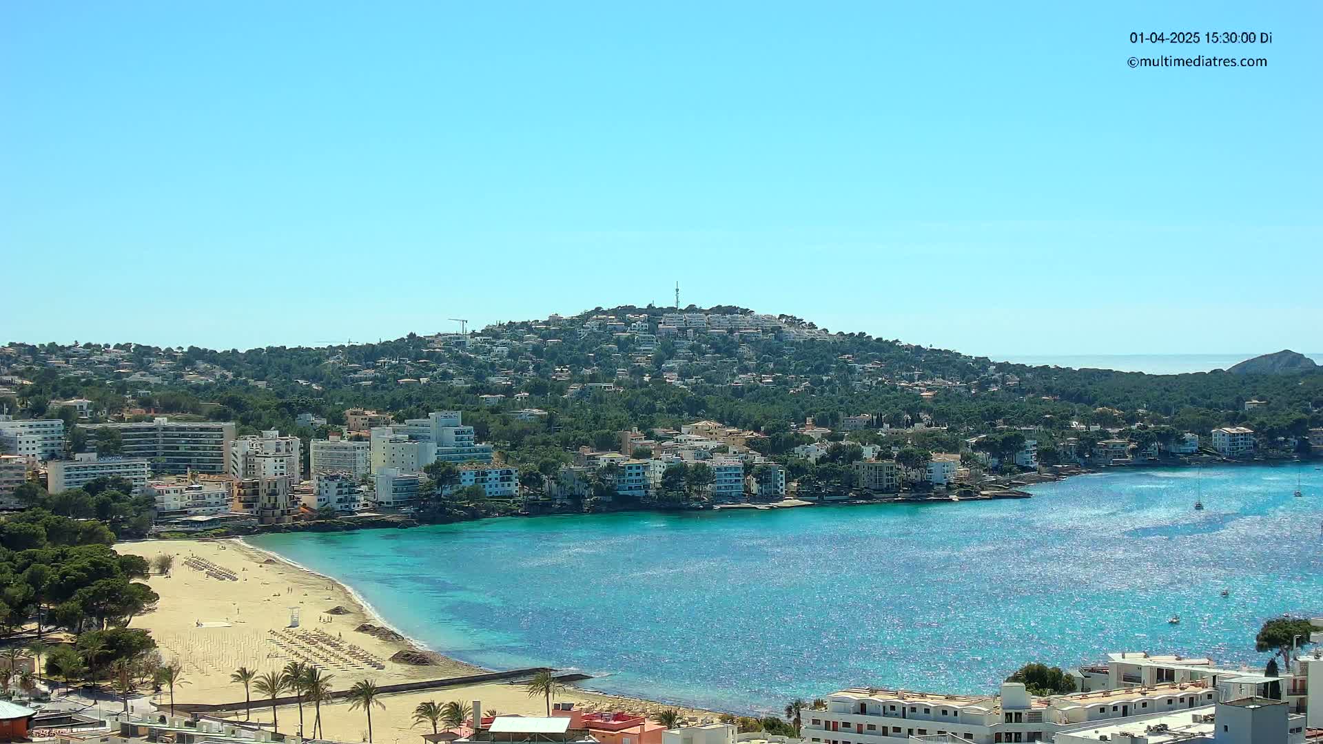 A clear, sunny day reveals a coastal town nestled against a hillside, with a sandy beach and calm, turquoise waters.