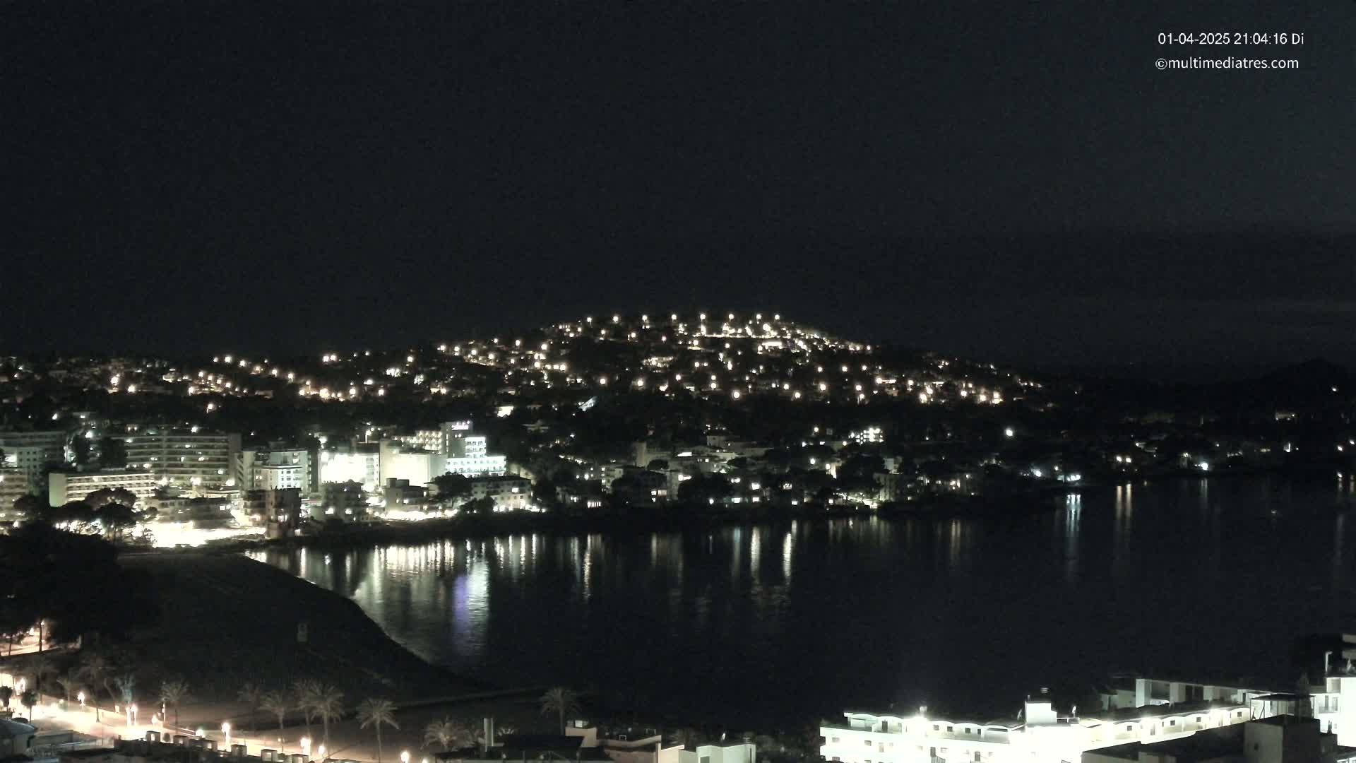A nighttime view of a town situated on a hill overlooking a calm body of water, with the lights of buildings and streetlights reflecting on the water's surface.