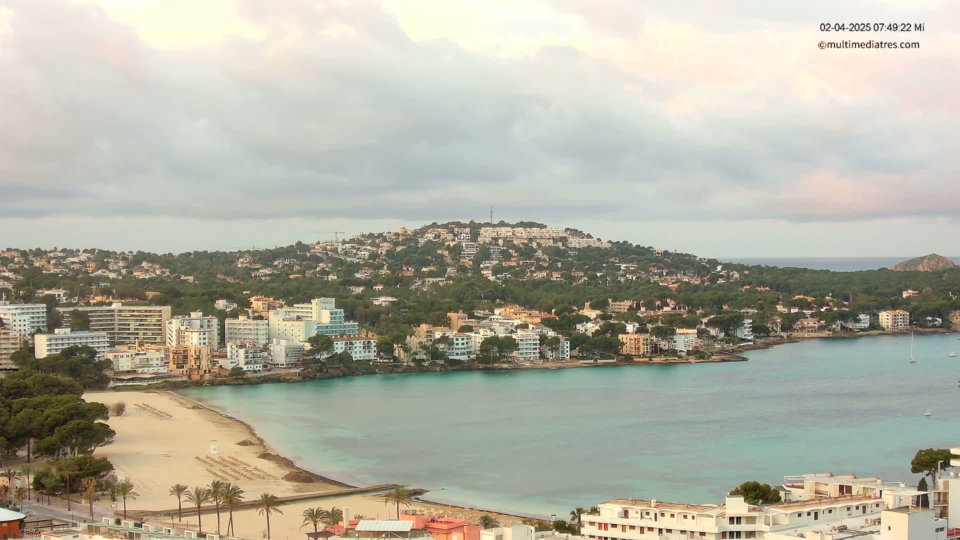 A coastal town with a sandy beach and turquoise water is nestled at the base of a hill covered in buildings and trees under a cloudy sky.