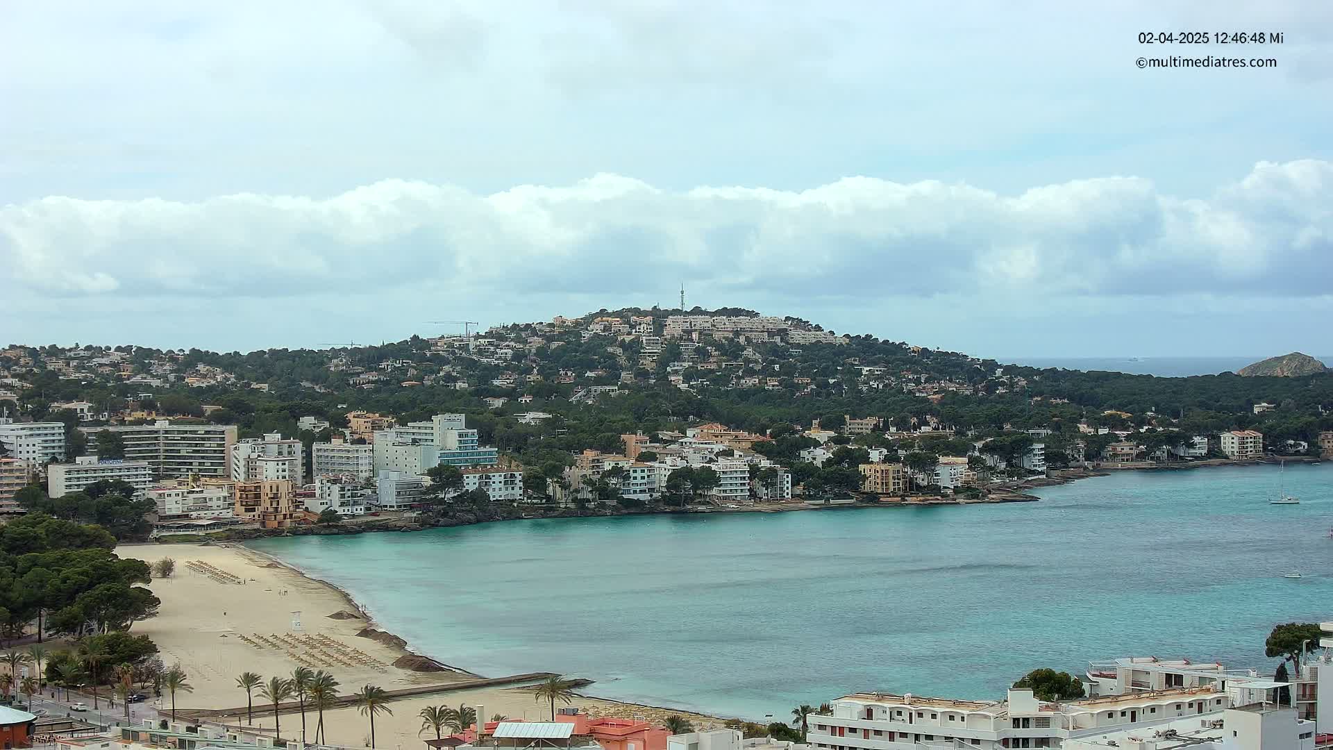 A coastal town with a sandy beach and turquoise water is nestled against a hill covered in buildings and vegetation under a partly cloudy sky.