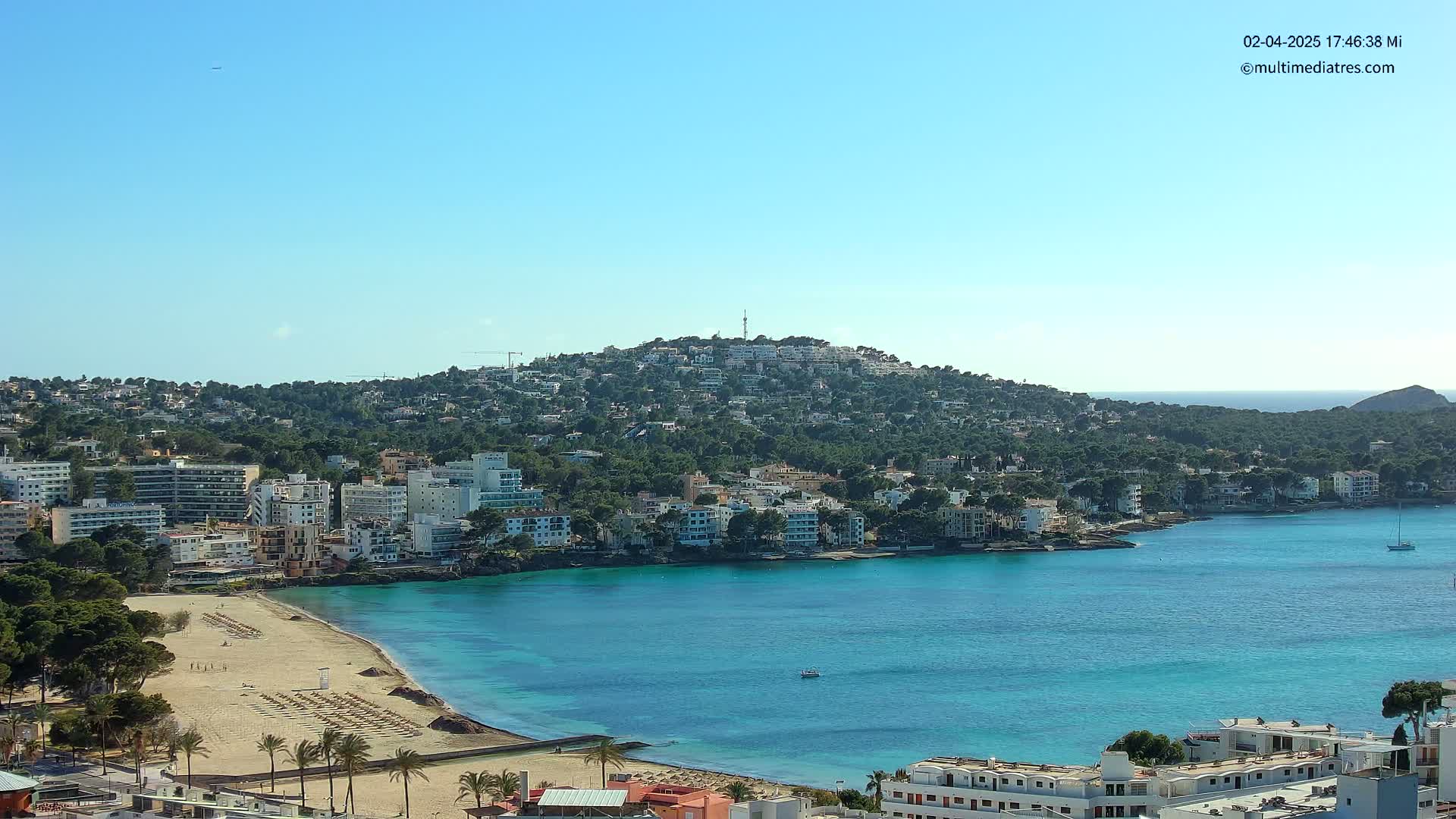 A hill covered in buildings overlooks a bay with a sandy beach and clear turquoise water under a bright blue sky.