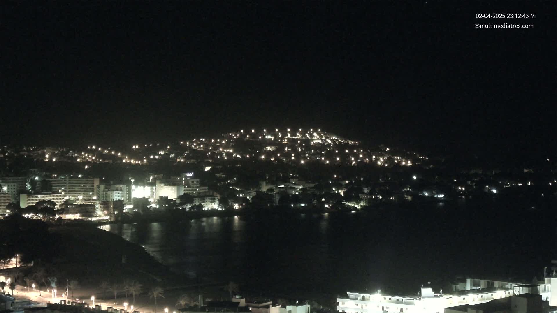 A nighttime view of a coastal town illuminated by many lights, with calm dark water in the foreground.