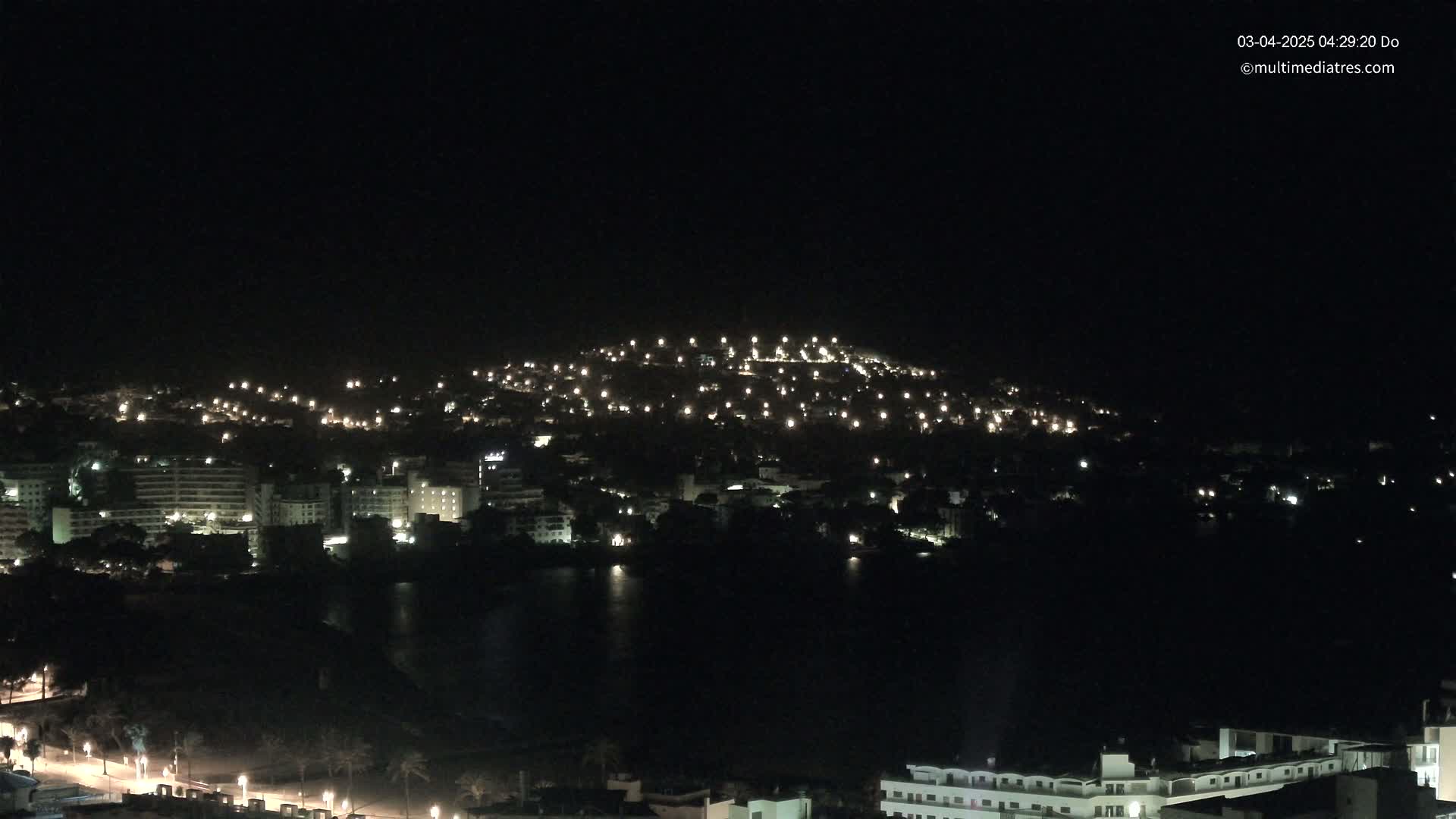 A nighttime view of a city on a hill overlooking a dark body of water, under a clear, dark sky.