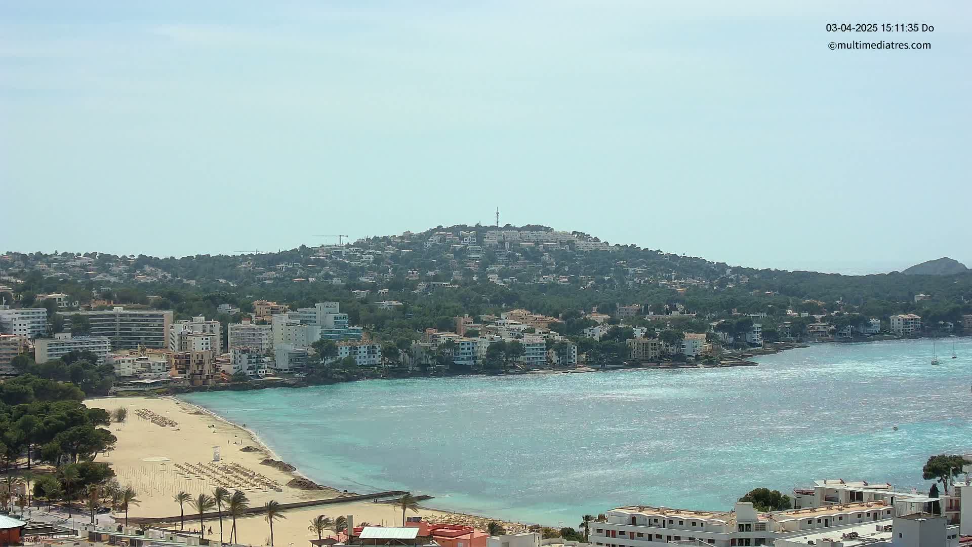 A coastal town with a sandy beach and turquoise water sits under a mostly clear sky, nestled below a hill covered in buildings and greenery.