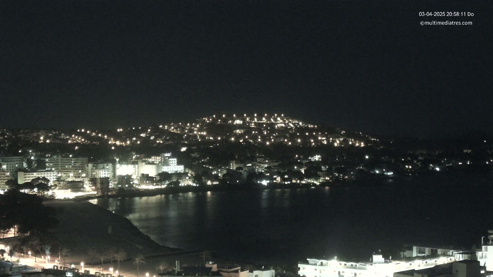 A nighttime view of a coastal town with buildings illuminated along a dark body of water, under a clear, dark sky.