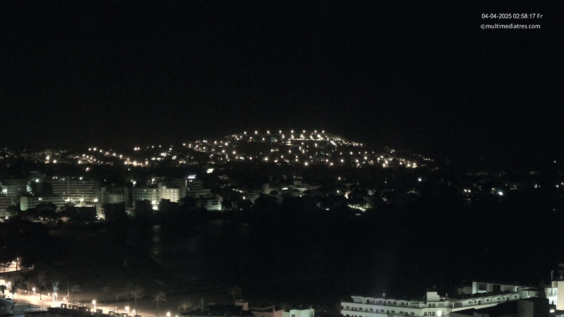A nighttime view of a city illuminated by numerous lights, nestled against a dark hill.