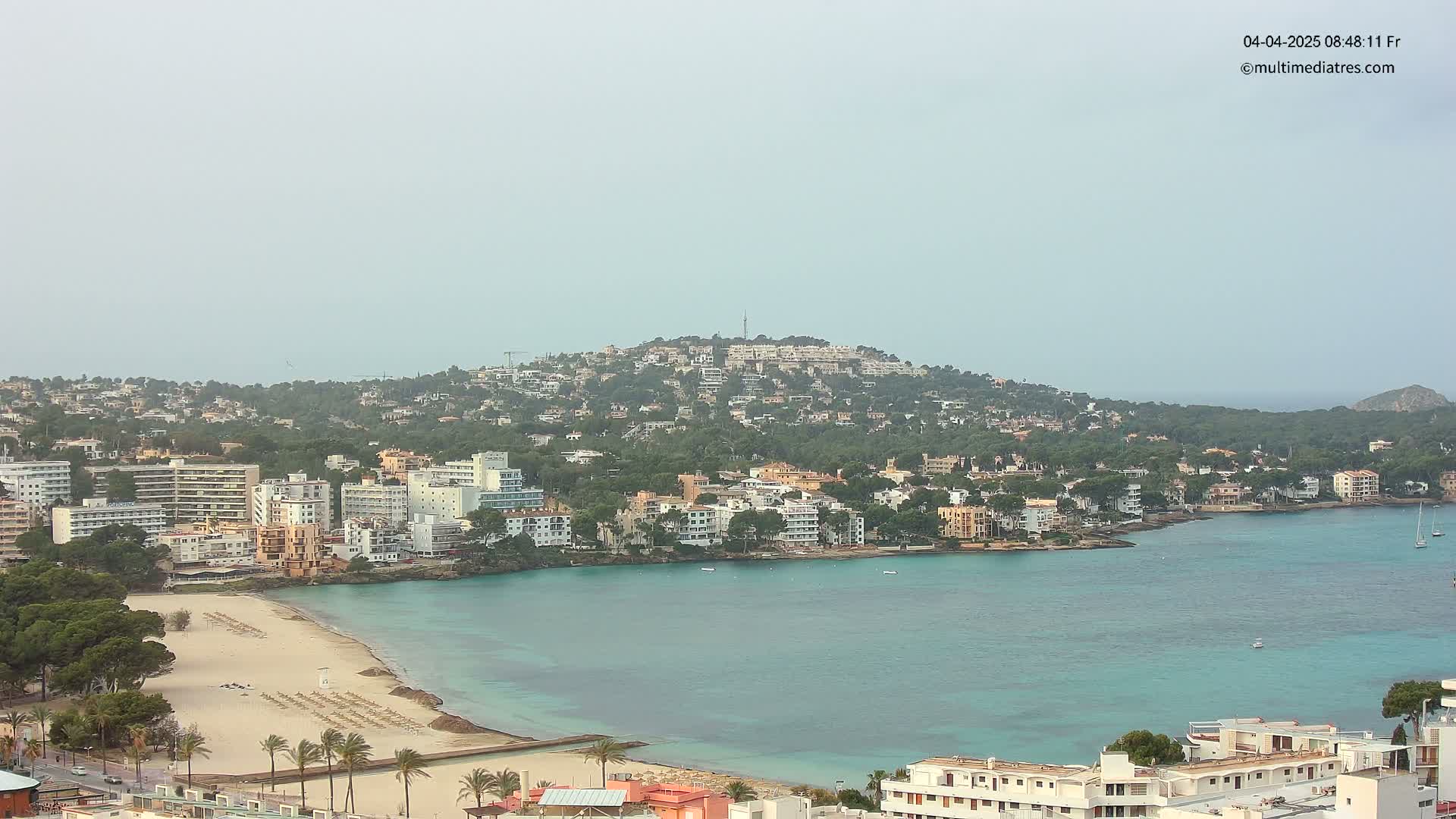 A hazy coastal town with a sandy beach and turquoise water is nestled against a hill covered in buildings and trees.