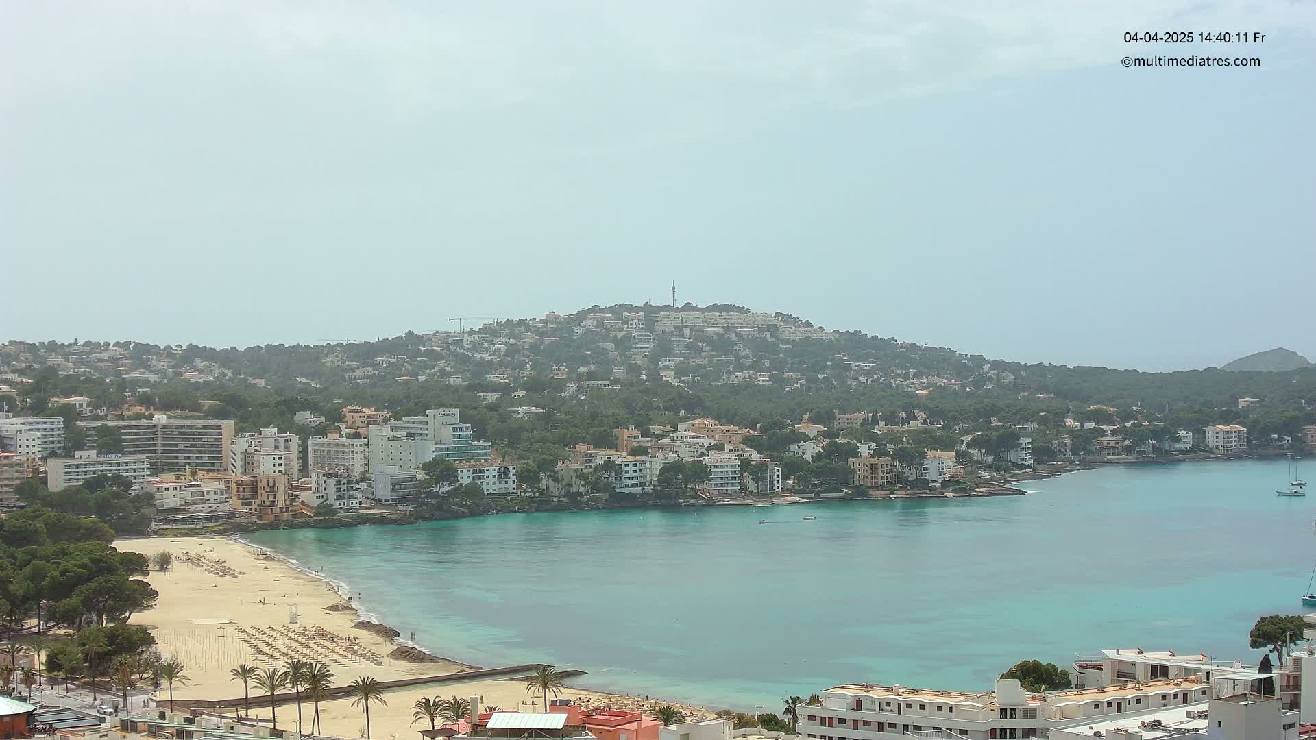 A hazy coastal town with a sandy beach and turquoise water nestled against a hill dotted with buildings.