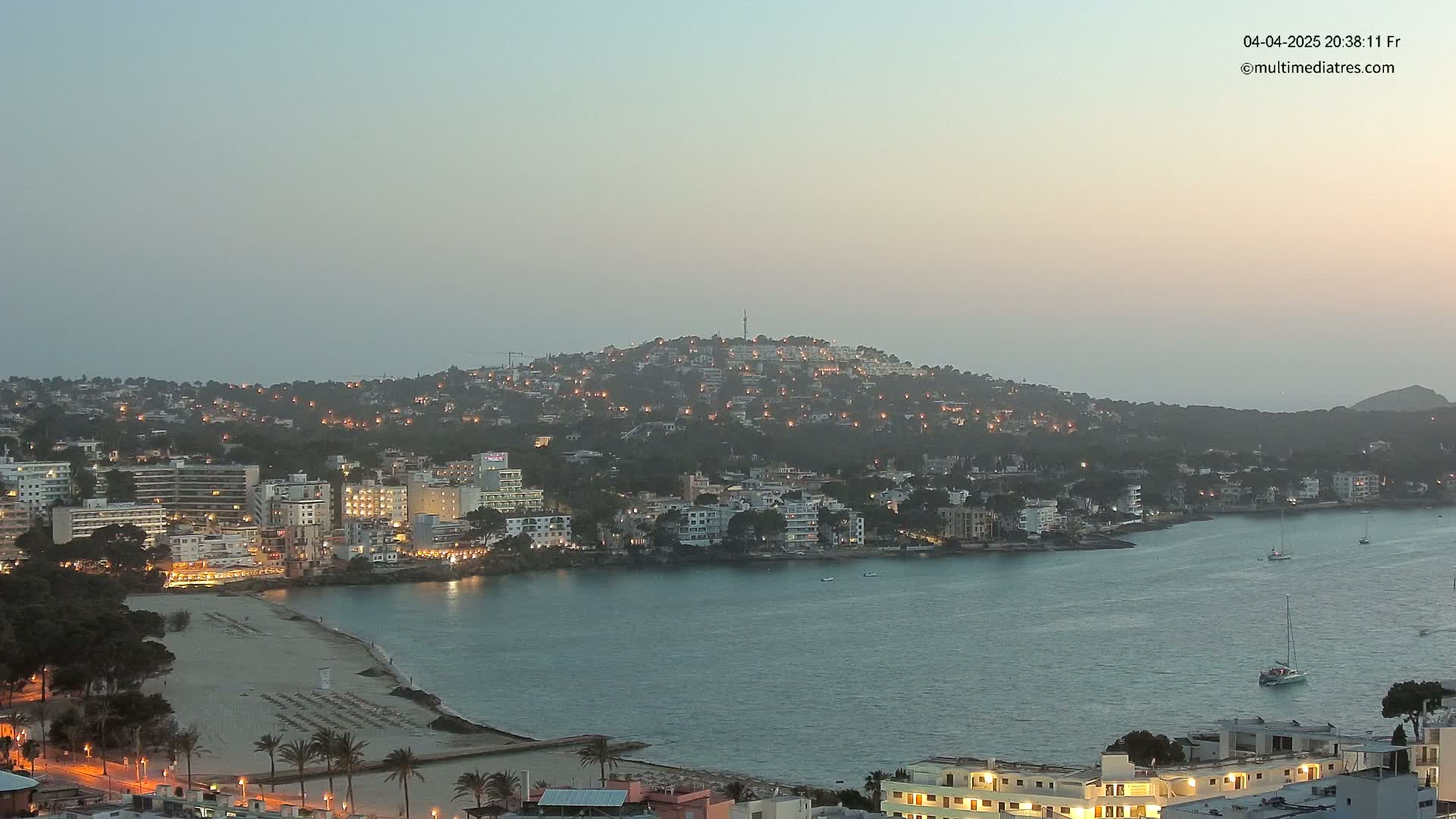 A coastal town at dusk, nestled on a hillside overlooking a calm bay with a few boats.