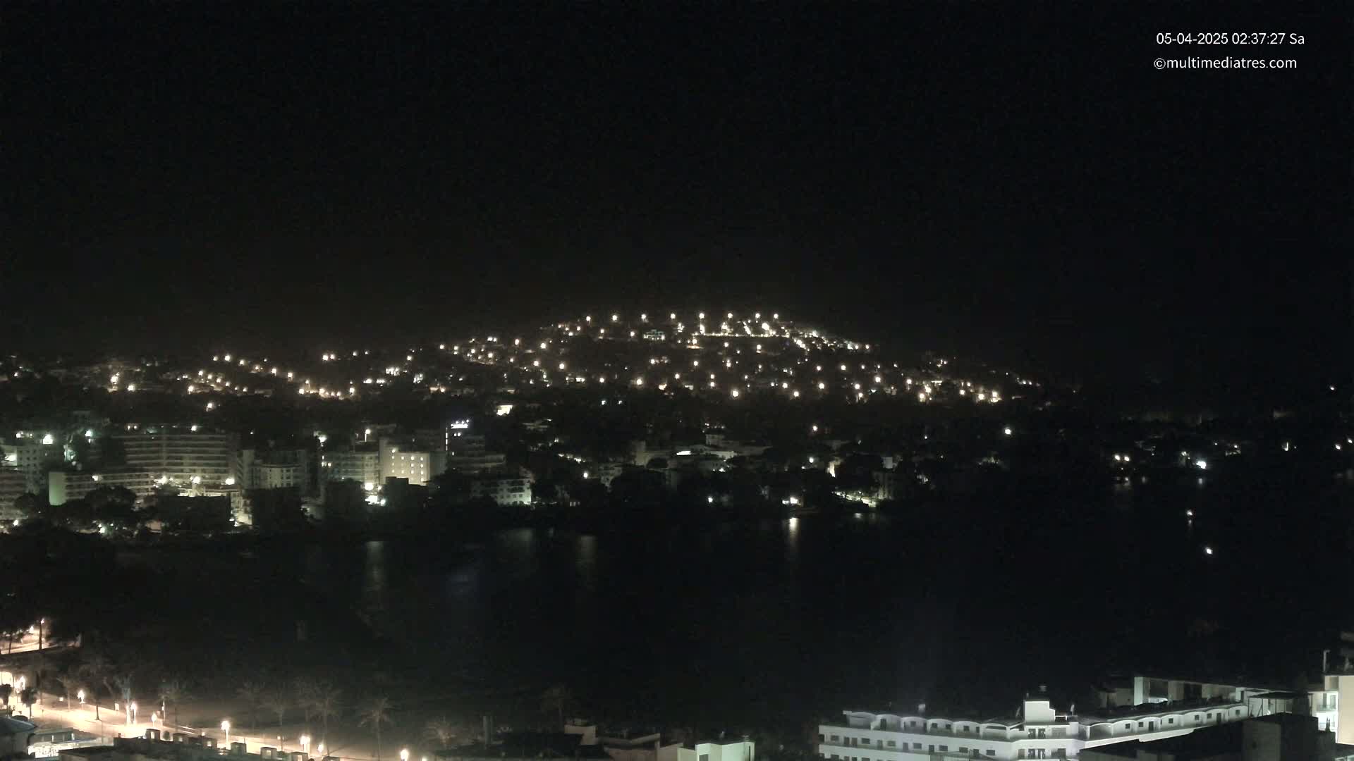 A nighttime view of a coastal city with numerous lights illuminating buildings and a hill, all reflected in a dark body of water under a clear, dark sky.