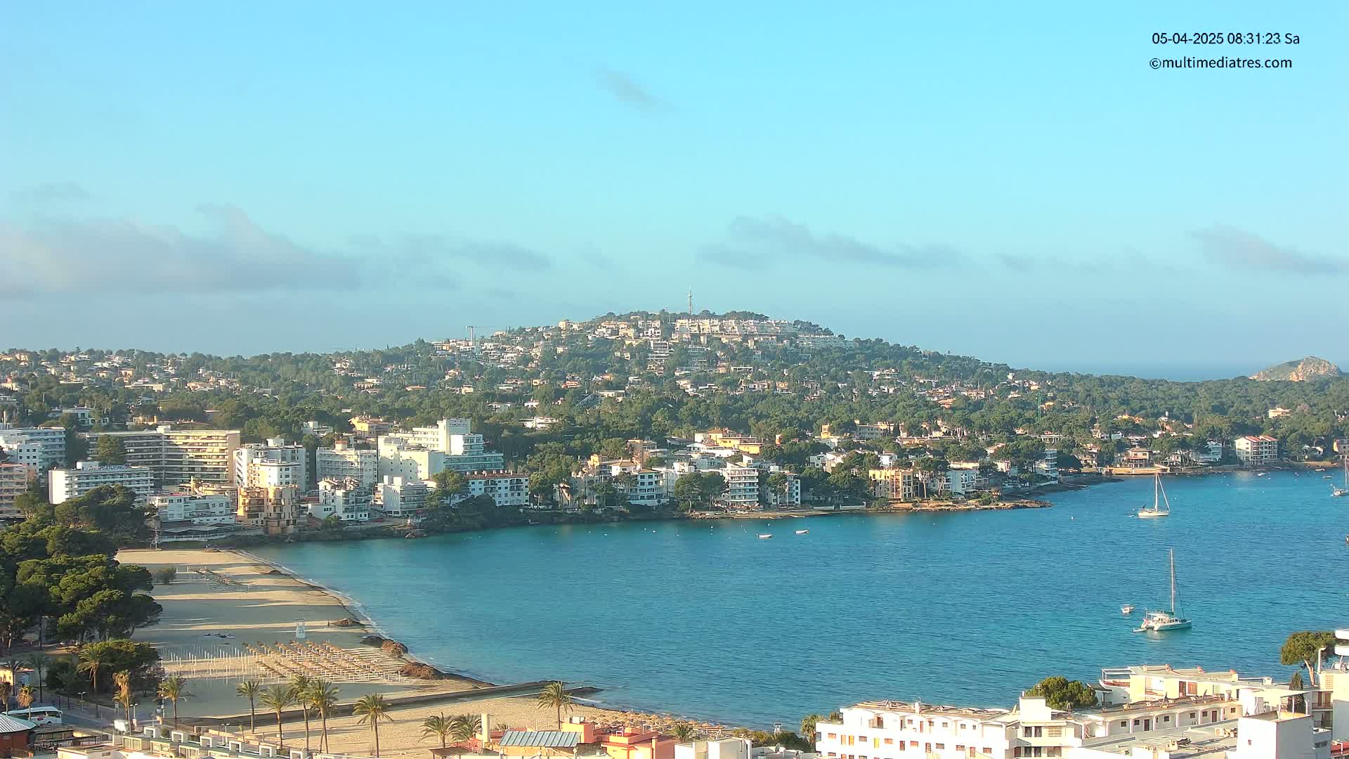 A coastal town with sandy beaches and turquoise waters under a mostly clear sky is nestled against a tree-covered hill.