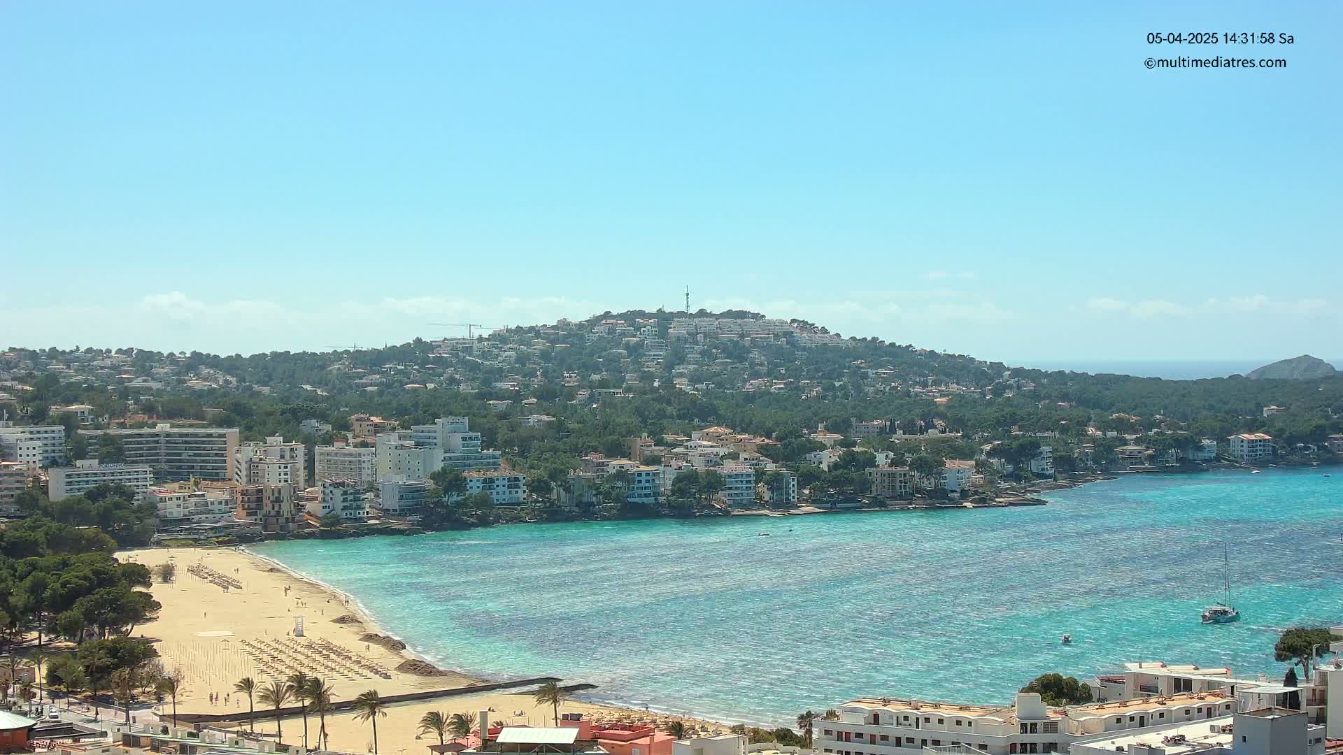 A sunny day reveals a coastal town nestled against a hill, featuring a sandy beach and turquoise waters.