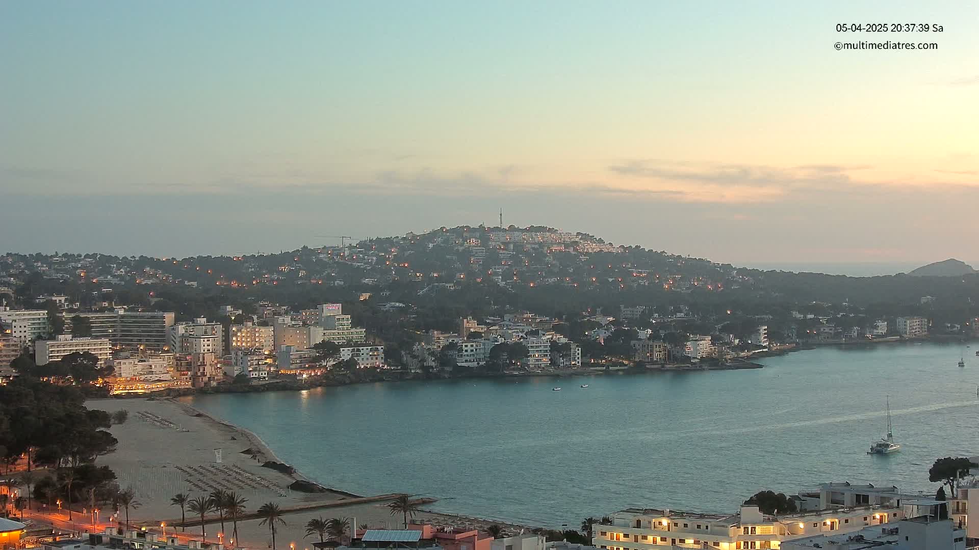 A coastal town at dusk, nestled against a hill dotted with lights, overlooks a calm bay with a sandy beach and a few boats.