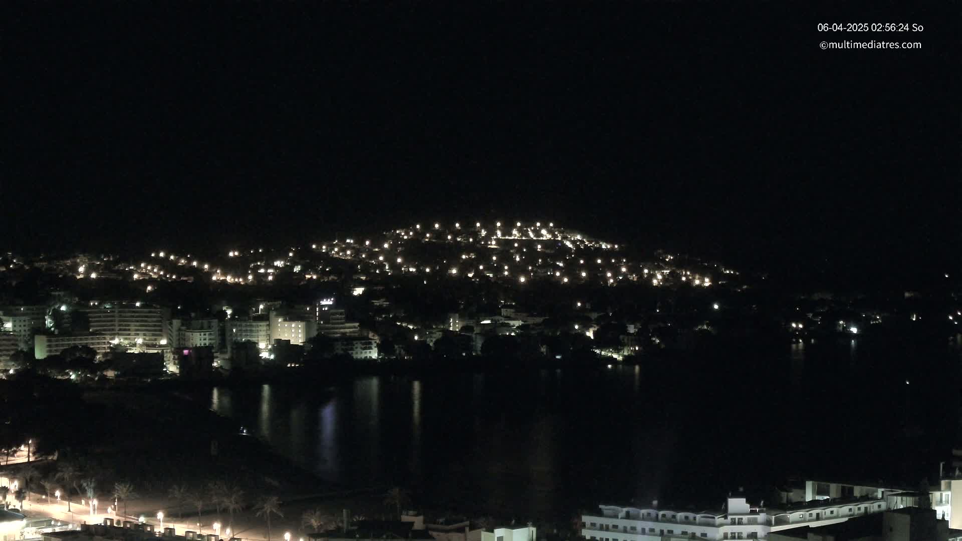 A nighttime view of a city illuminated by many lights, situated beside a dark body of water under a clear, dark sky.