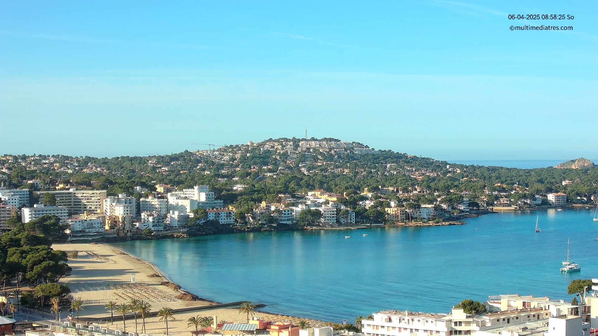 A coastal town with a sandy beach and clear blue water is nestled at the base of a hill covered in buildings and trees under a bright, sunny sky.