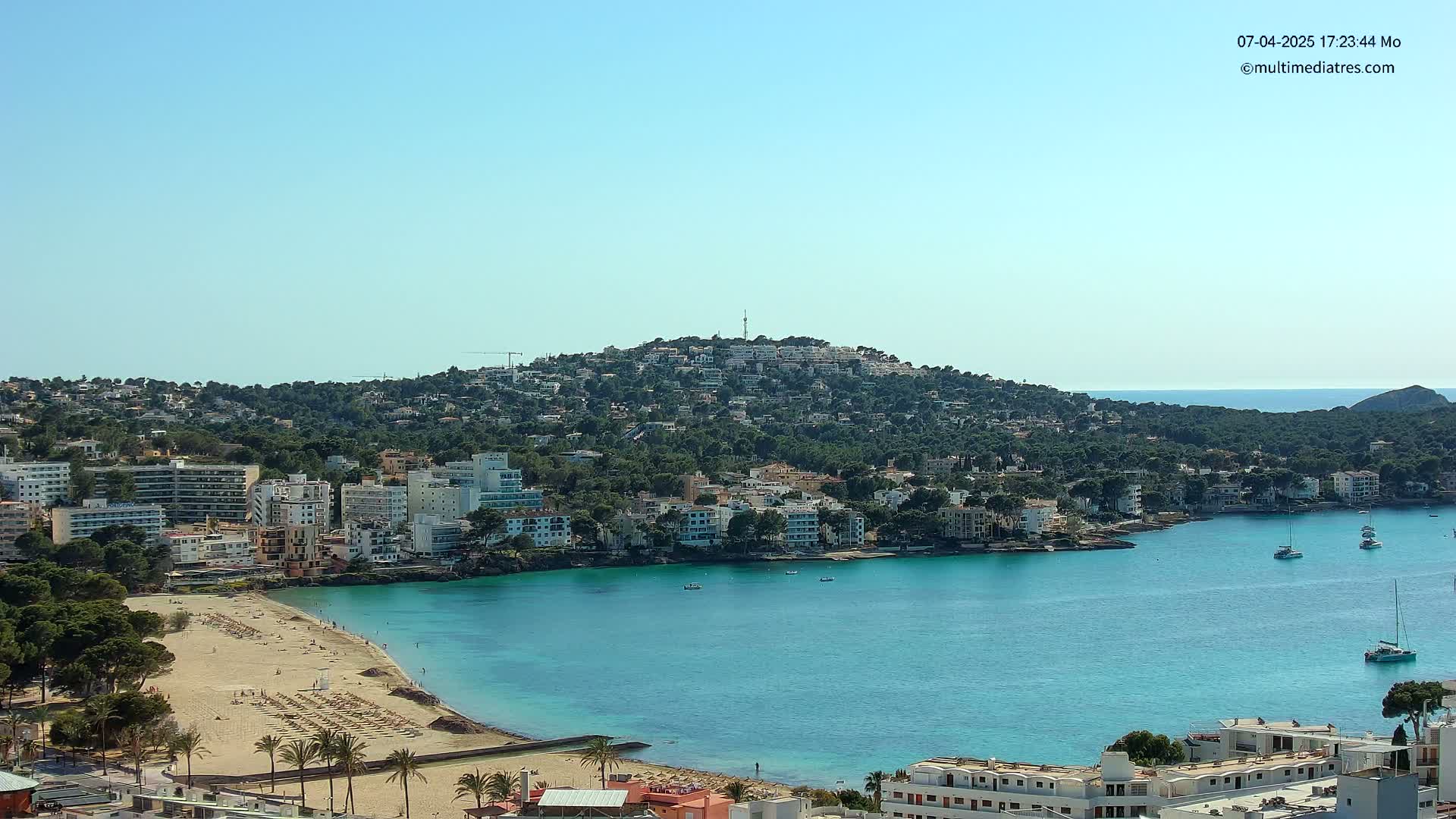 A coastal town with a sandy beach and turquoise water sits nestled below a hill covered in buildings and lush green vegetation under a clear blue sky.