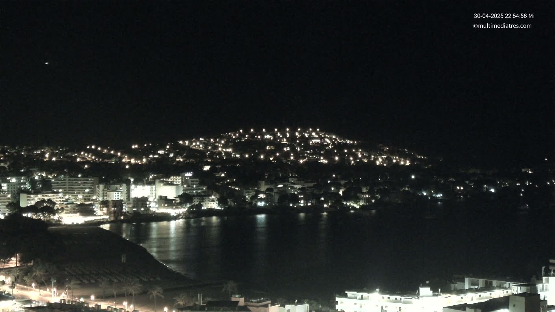 A nighttime view of a coastal town illuminated by many lights, with a dark body of water in the foreground under a clear, dark sky.