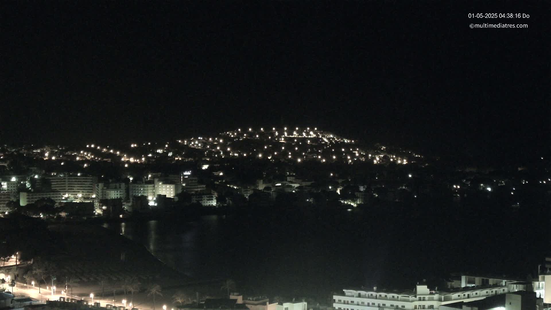 A nighttime view of a city nestled on a hill, illuminated by numerous lights, with a dark body of water in the foreground.