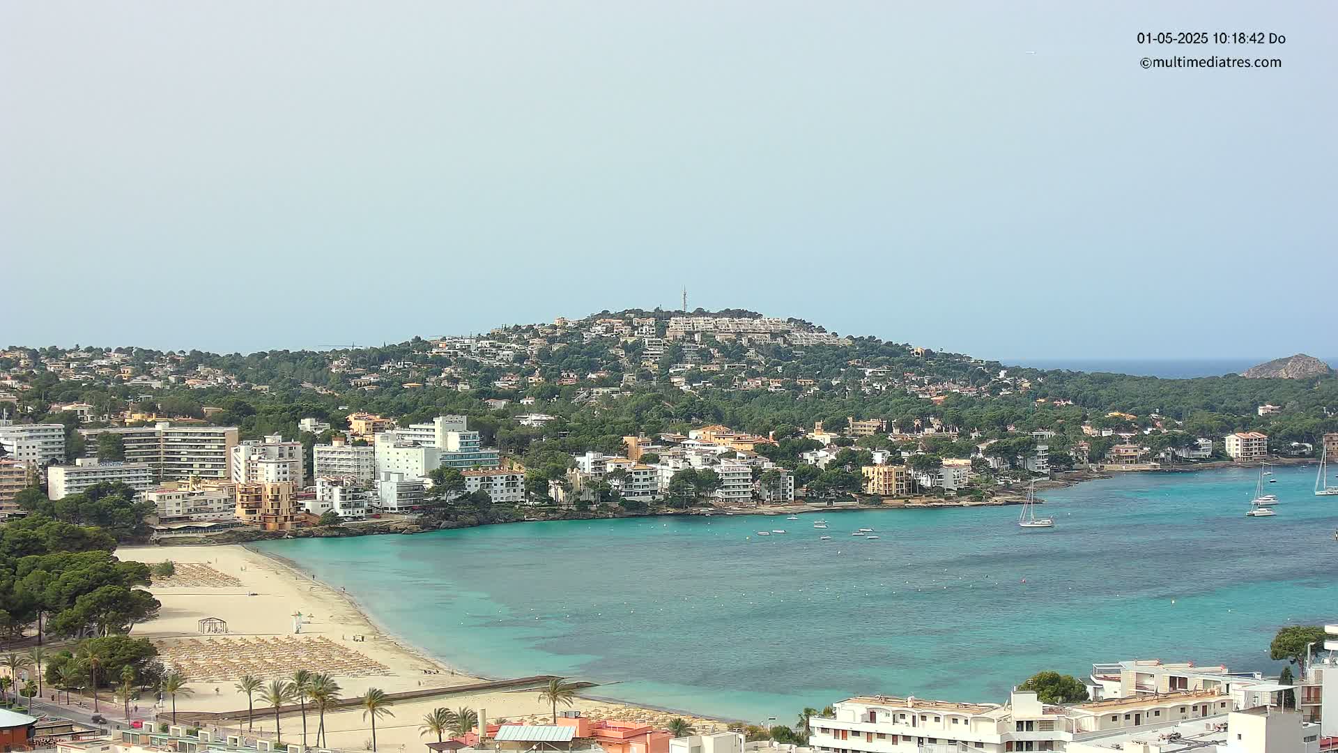 A coastal town with numerous buildings hugs a calm, turquoise bay under a hazy sky, with a hill covered in buildings and trees in the background.