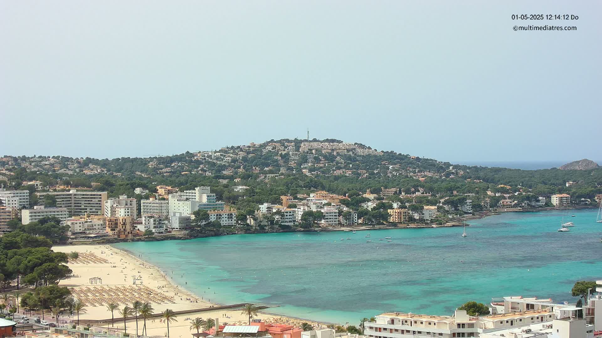 A coastal town with turquoise water, sandy beach, and numerous buildings sprawls across a hillside under a clear sky.