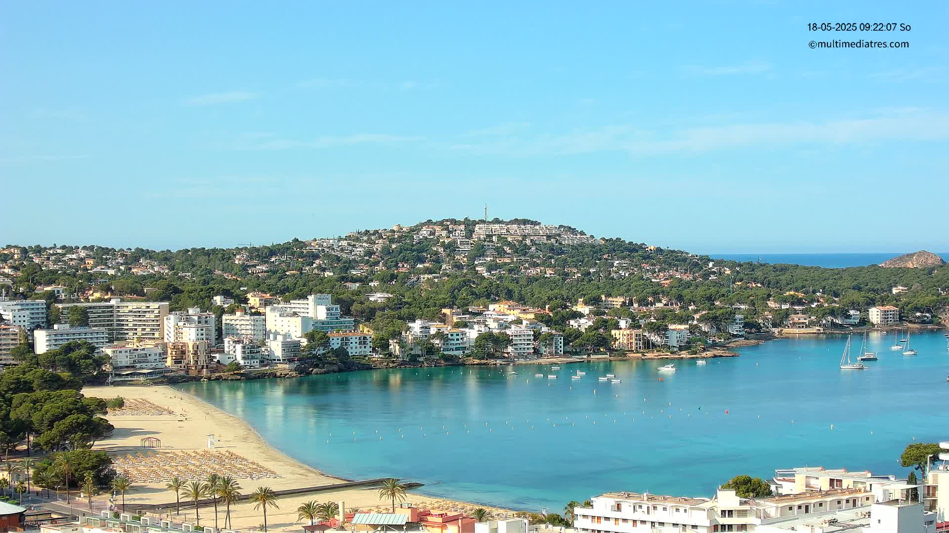 A sandy beach with lounge chairs curves into a calm, turquoise bay filled with boats, surrounded by a hillside town under a clear blue sky.