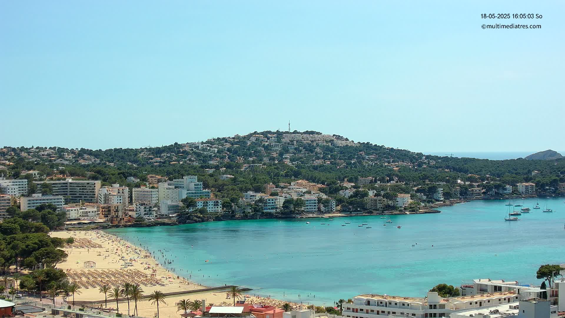 A sunny day reveals a coastal town nestled beside a turquoise bay, with a hill covered in buildings and lush greenery in the background.