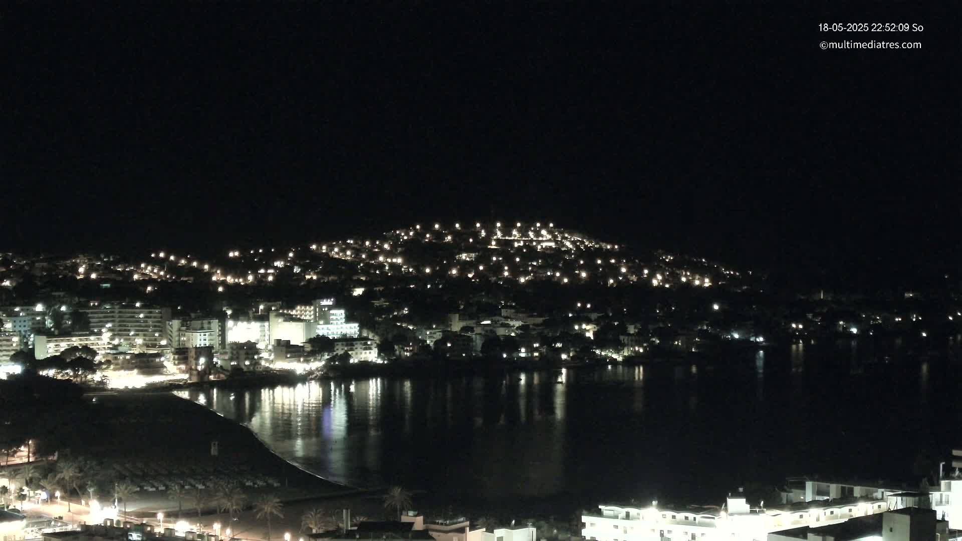 A hilltop town is brightly lit at night, its lights reflected in the calm waters of a bay below.