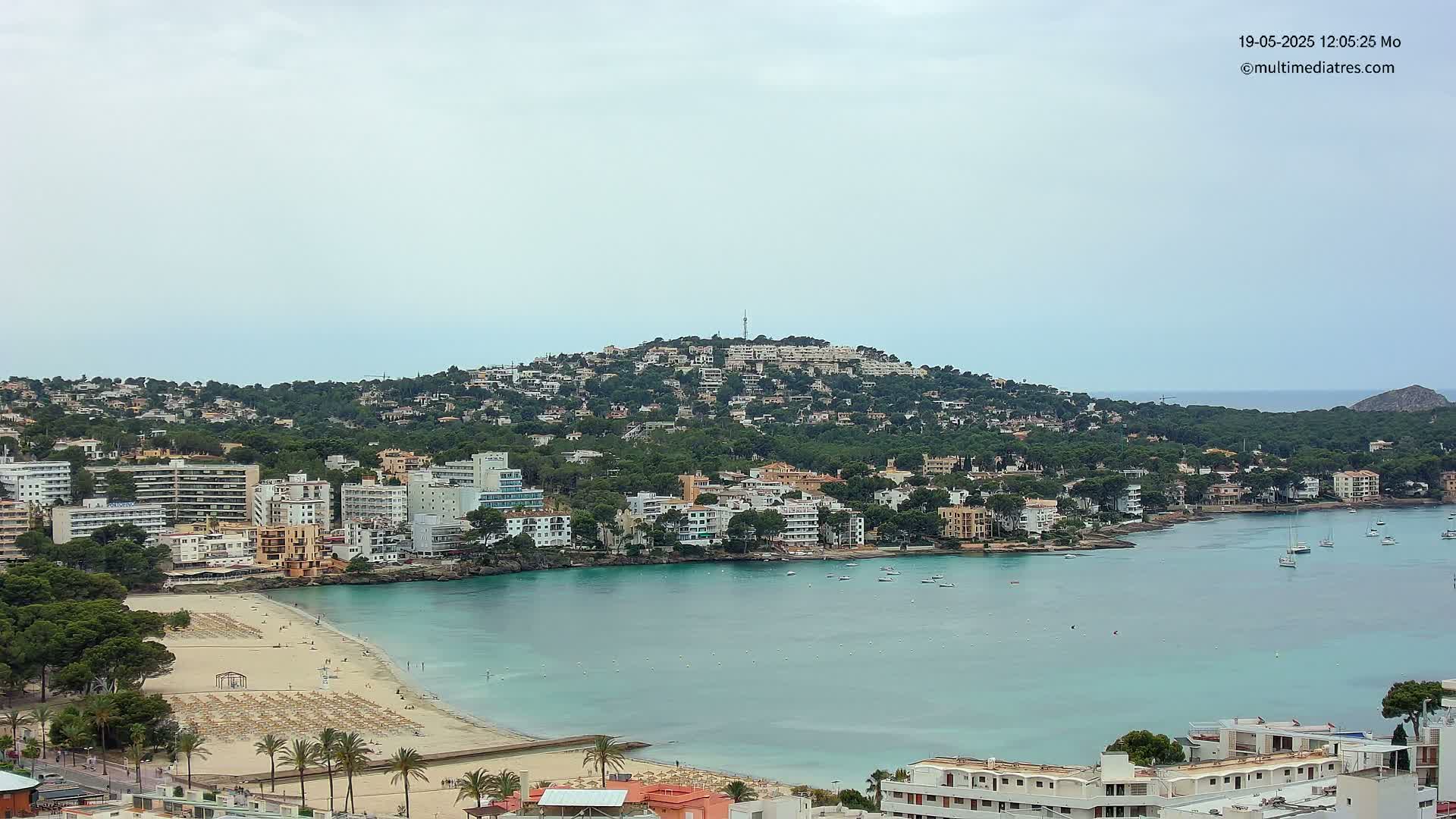 A coastal town with a sandy beach and calm turquoise water sits nestled below a hill covered in buildings and lush green vegetation under a mostly cloudy sky.