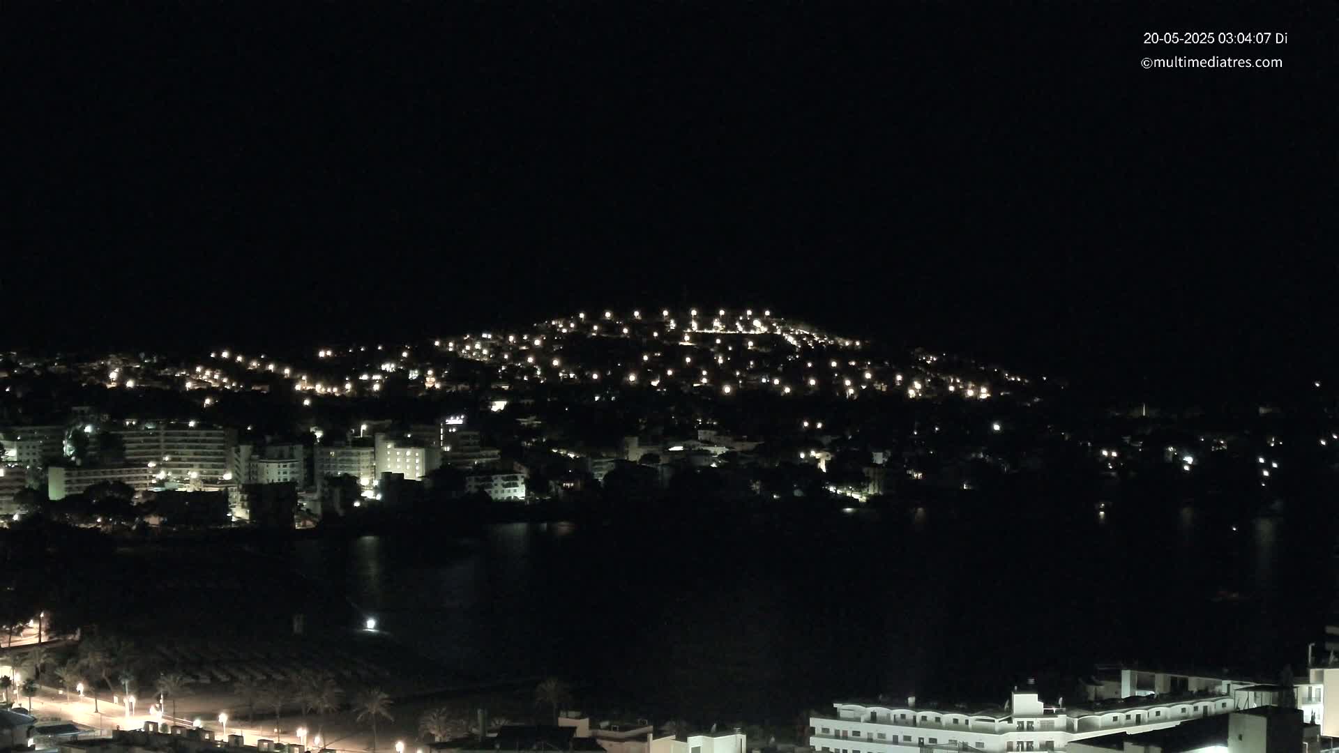 A nighttime view of a coastal town with many buildings illuminated, showing a dark body of water in the foreground.