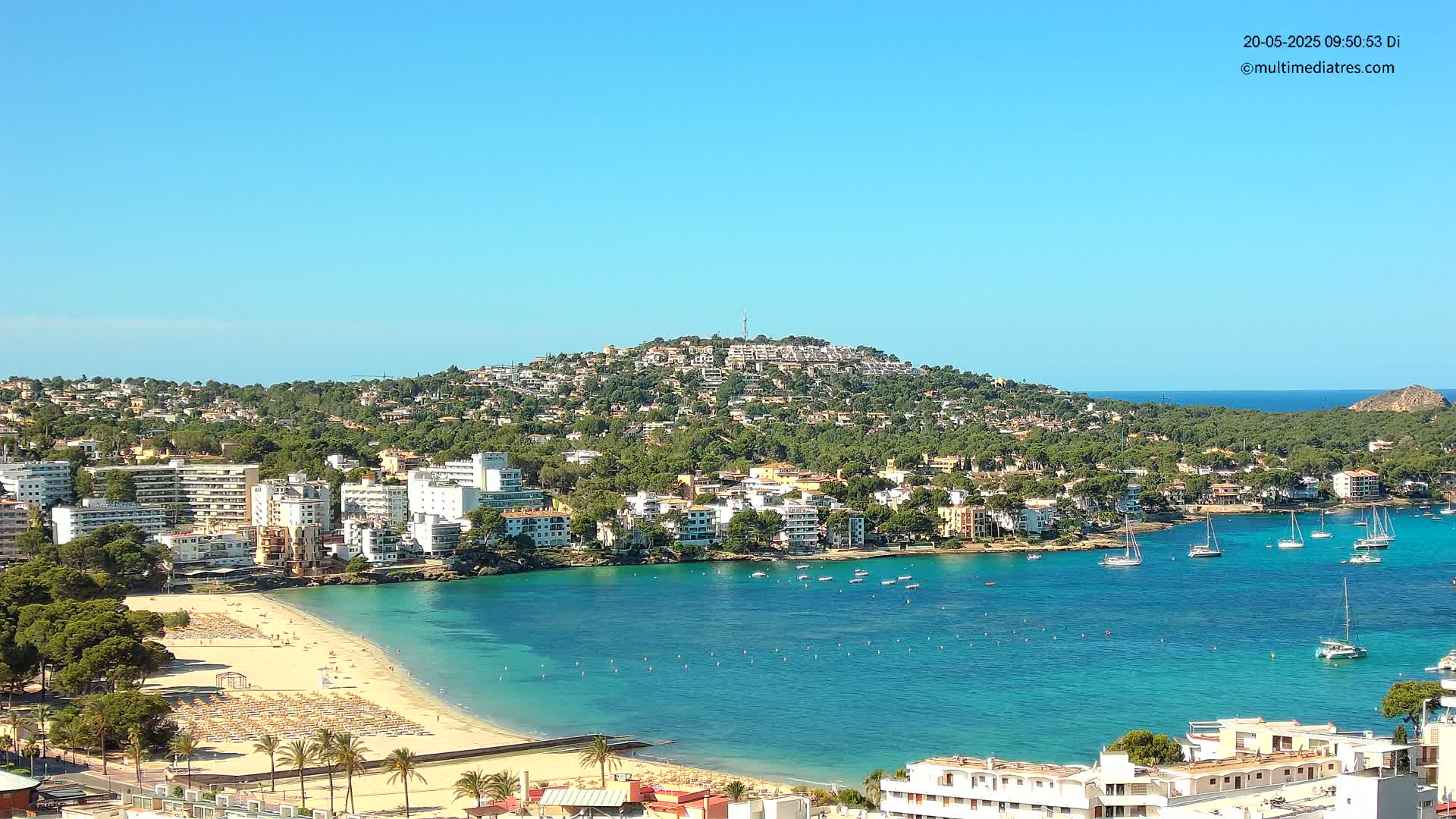 A coastal town with a sandy beach and turquoise water sits beneath a hill covered in buildings and trees on a clear, sunny day.