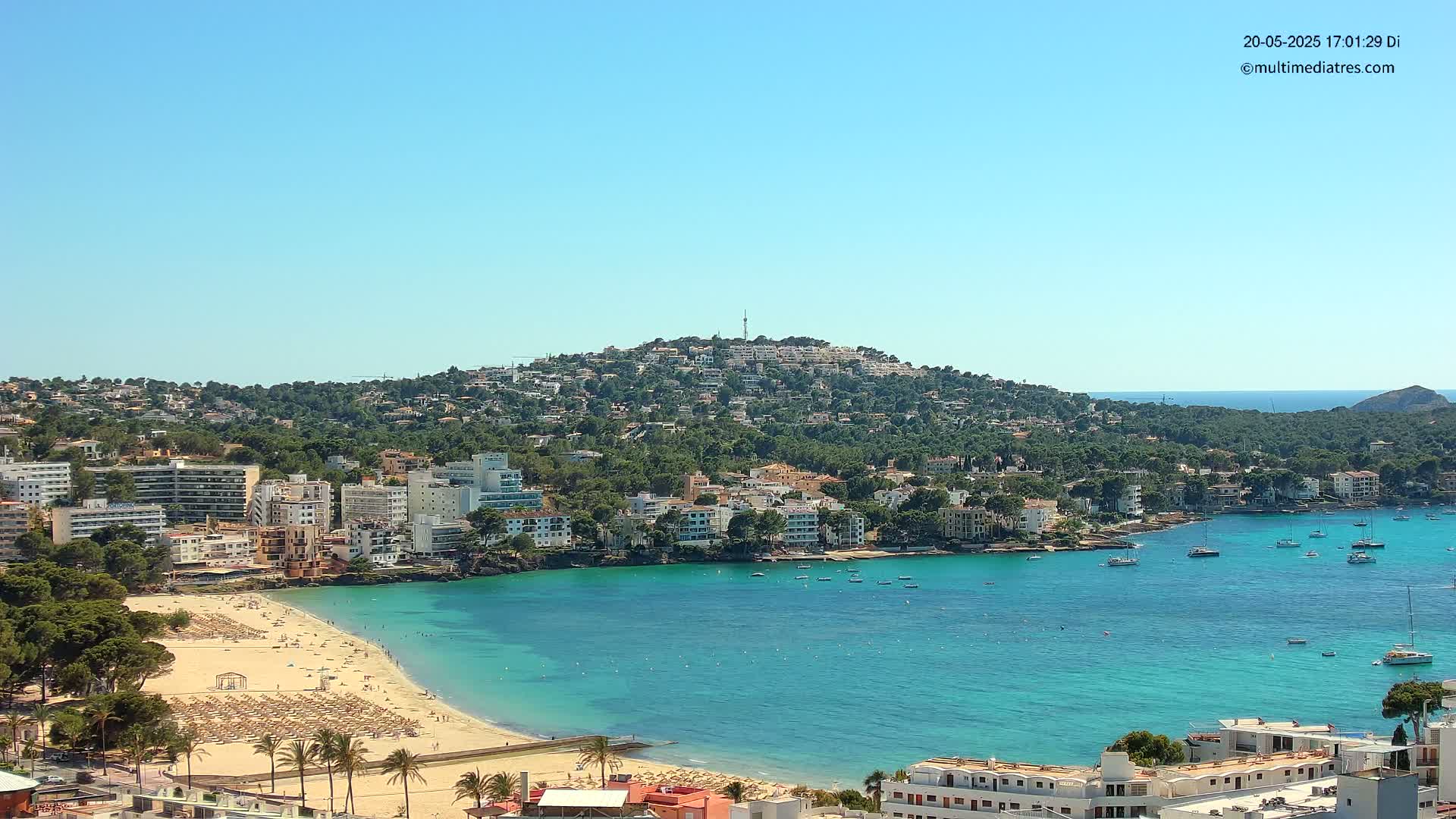 A sunny coastal town with a sandy beach, turquoise water, and numerous boats in a calm bay, nestled at the base of a hill covered in buildings and greenery.