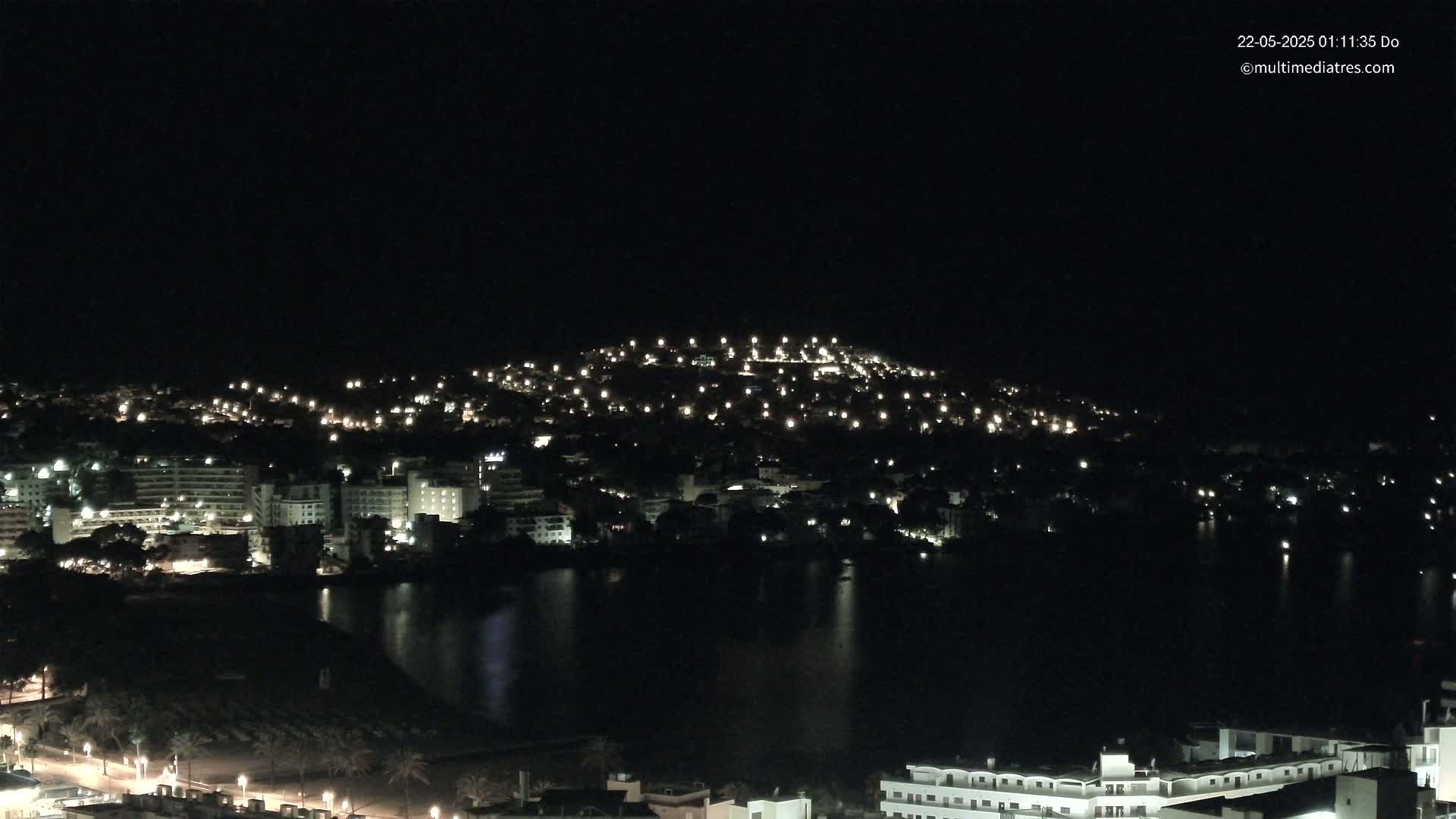 A nighttime view of a coastal town with numerous lights illuminating buildings and a hill, reflected in a dark body of water under a clear sky.