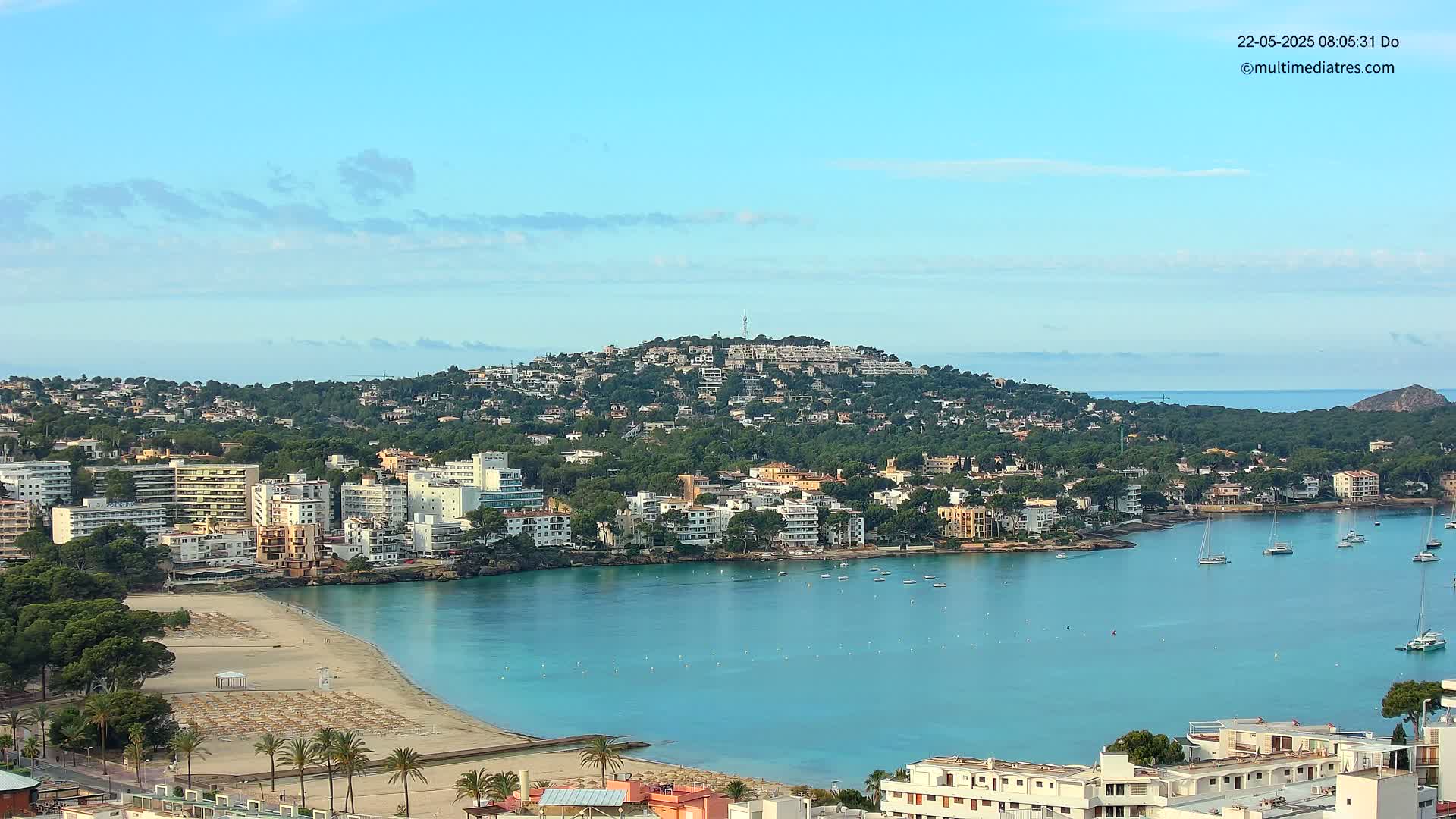 A coastal town nestled against a verdant hill overlooking a calm, turquoise bay under a clear, light-blue sky.