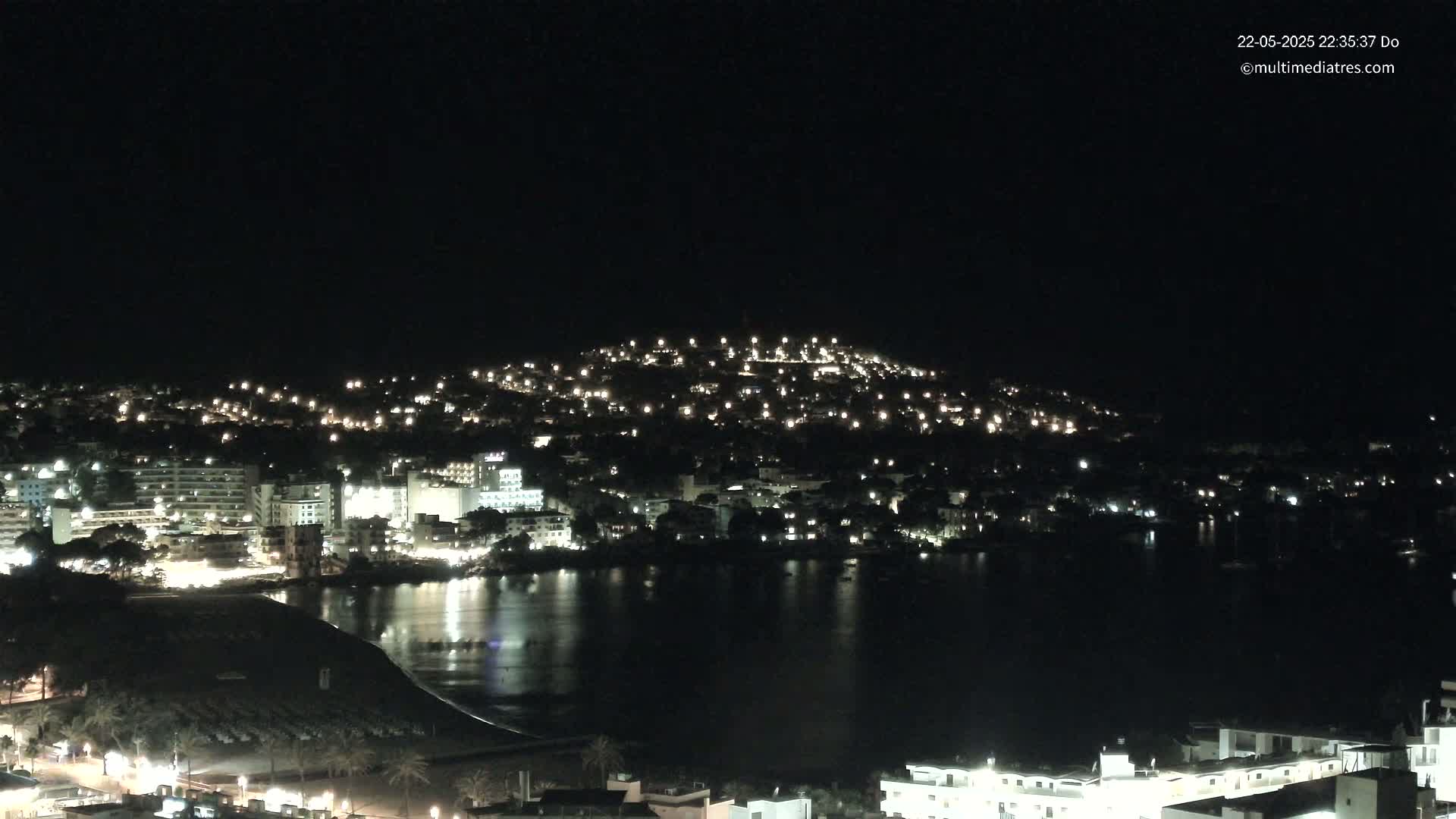 A nighttime view of a coastal town illuminated by many lights, with calm water reflecting the lights.