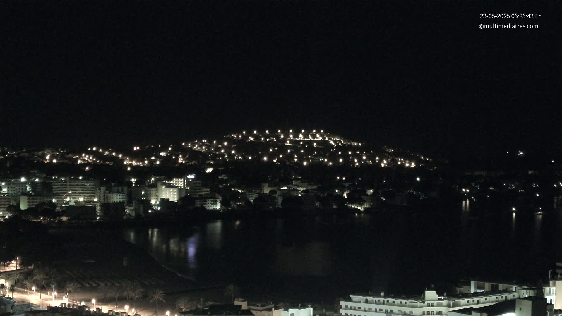 A nighttime view of a coastal town with numerous lights illuminating buildings and streets, nestled around a dark body of water under a clear sky.