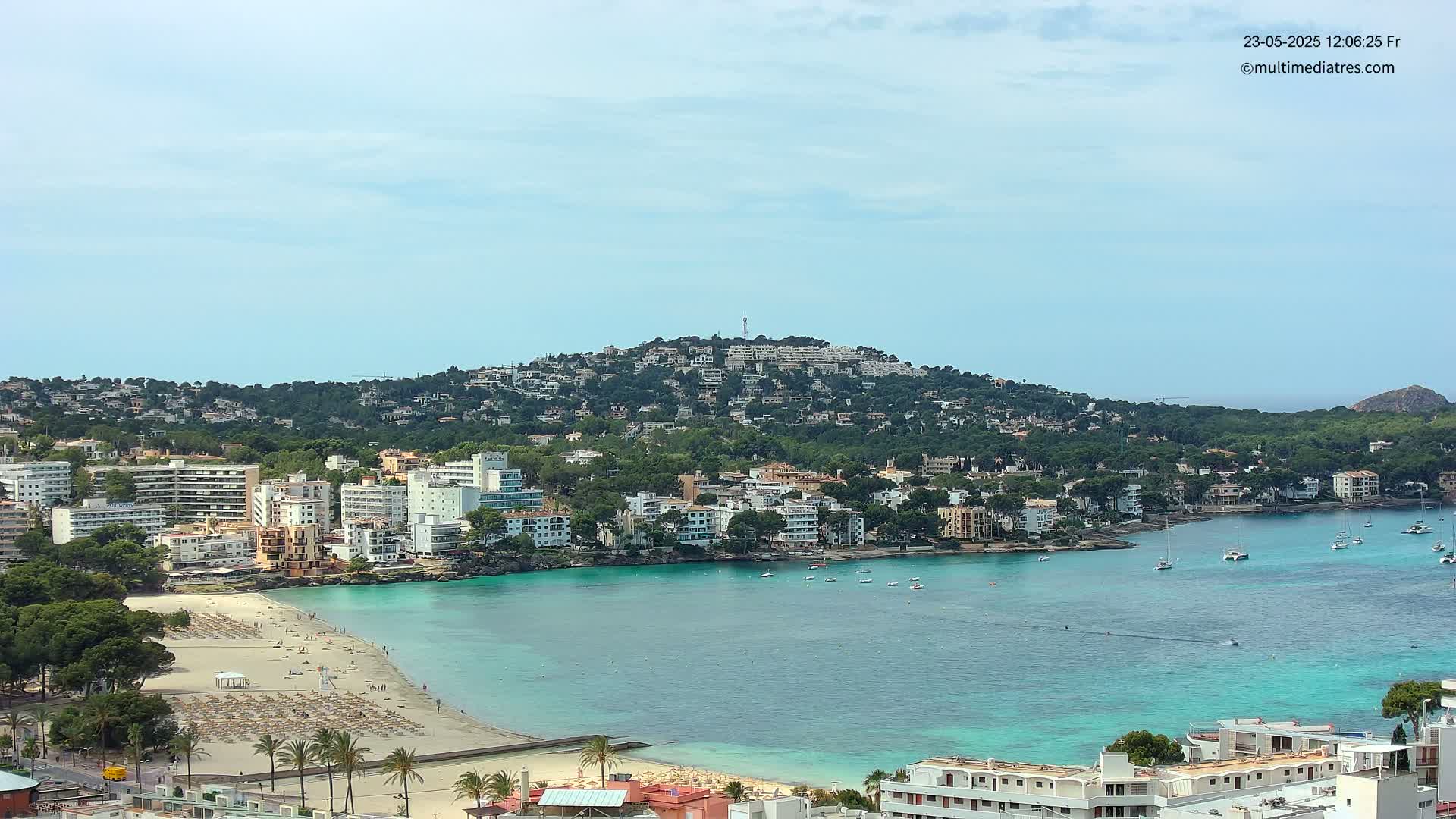 A coastal town with a sandy beach and turquoise water sits below a hill covered in buildings and trees under a mostly clear sky.