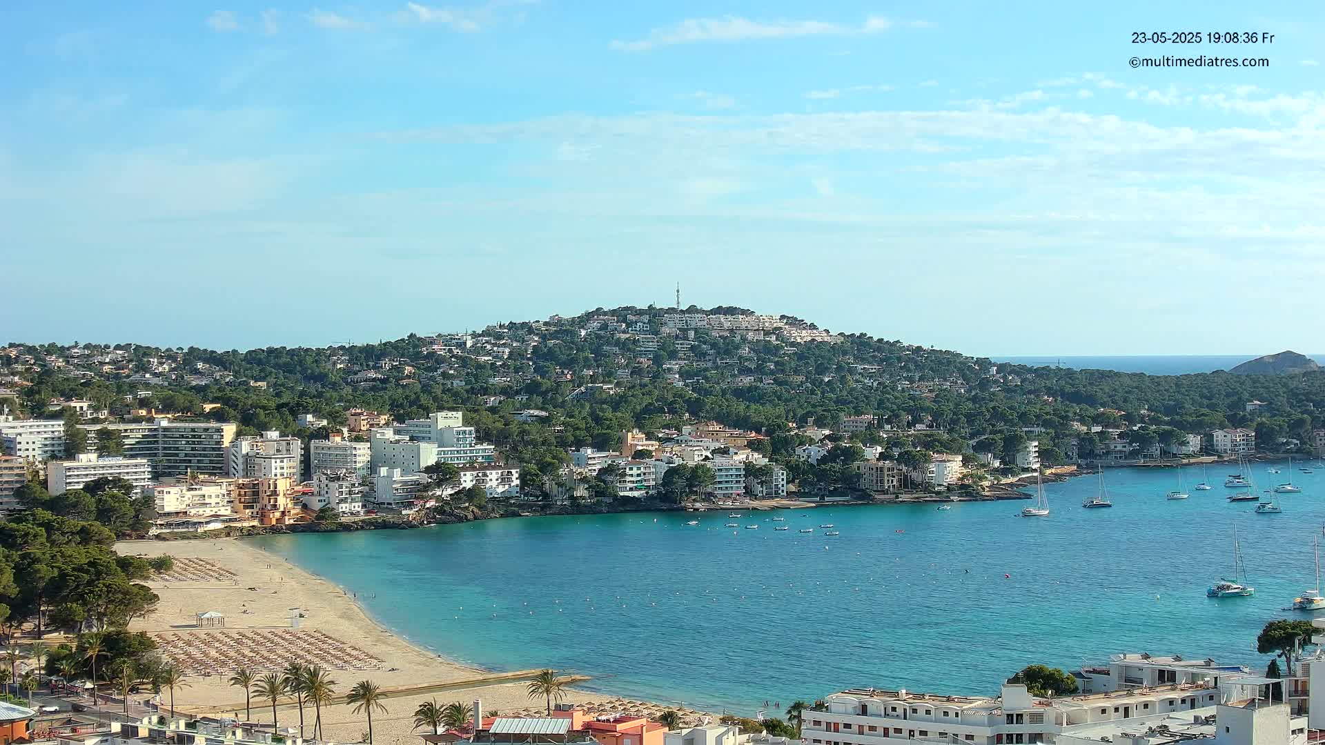 A coastal town nestled against a green hill, with a sandy beach and calm, turquoise water dotted with boats under a mostly clear, sunny sky.
