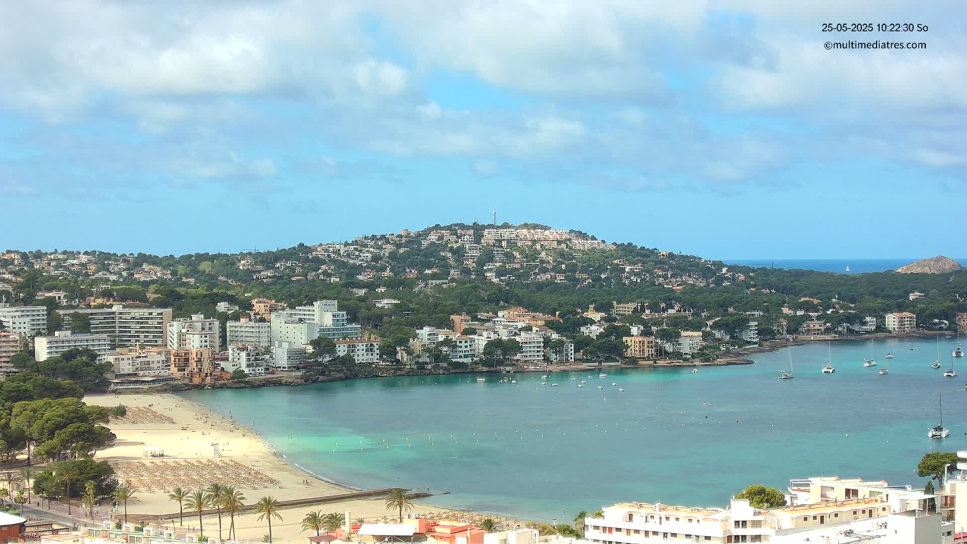 A coastal town with a sandy beach and turquoise water is nestled below a hill covered in buildings and trees under a partly cloudy sky.